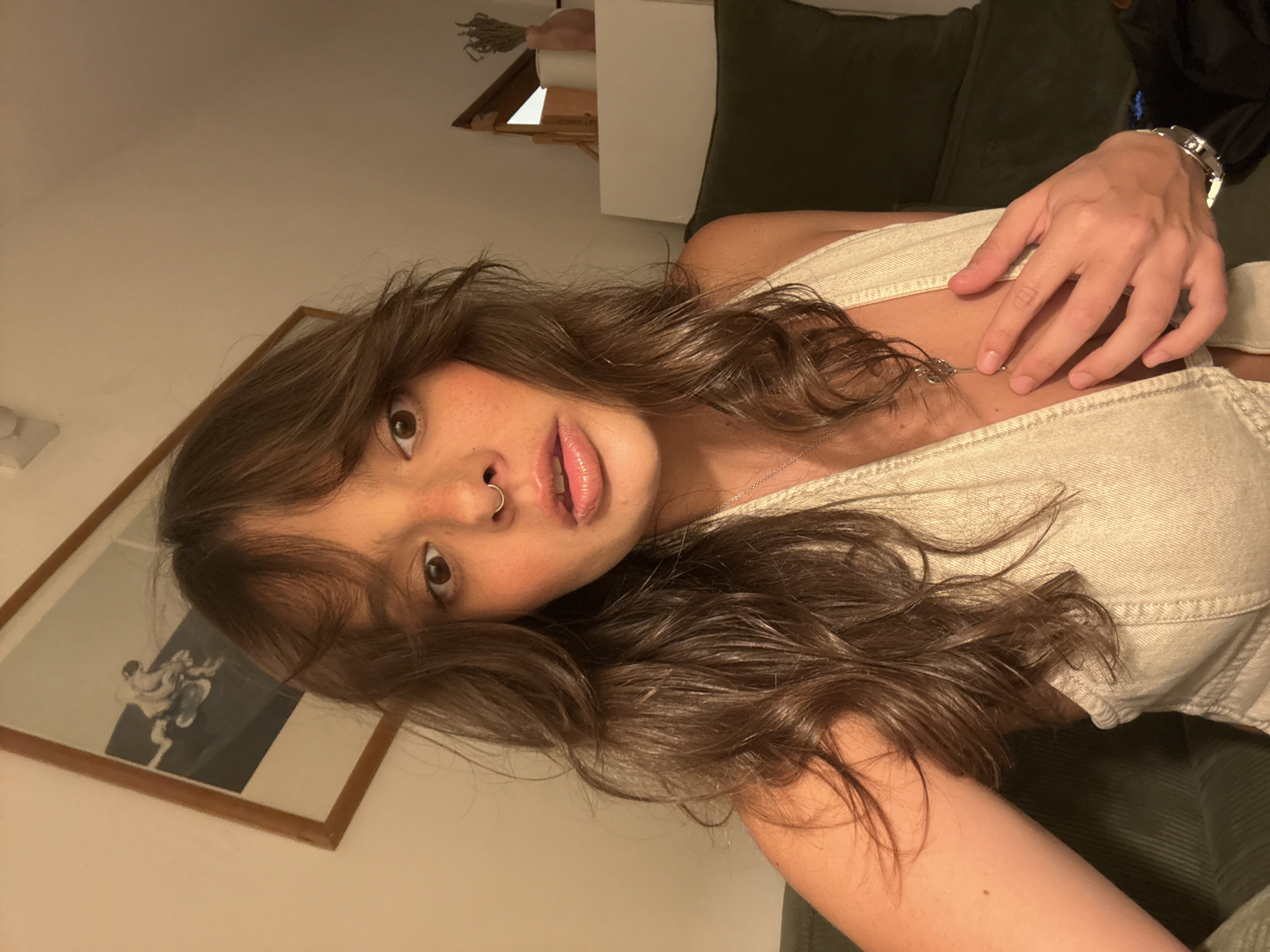 A young woman with long brown hair and a nose ring is looking at the camera. She is wearing a light-colored denim top and a silver necklace. A hand with a silver watch is resting on her chest. She is sitting on a dark green corduroy couch. A framed picture hangs on the wall behind her, and a white dresser is visible in the background. The image was likely taken indoors. The location in Tel-Aviv, Israel is unknown.