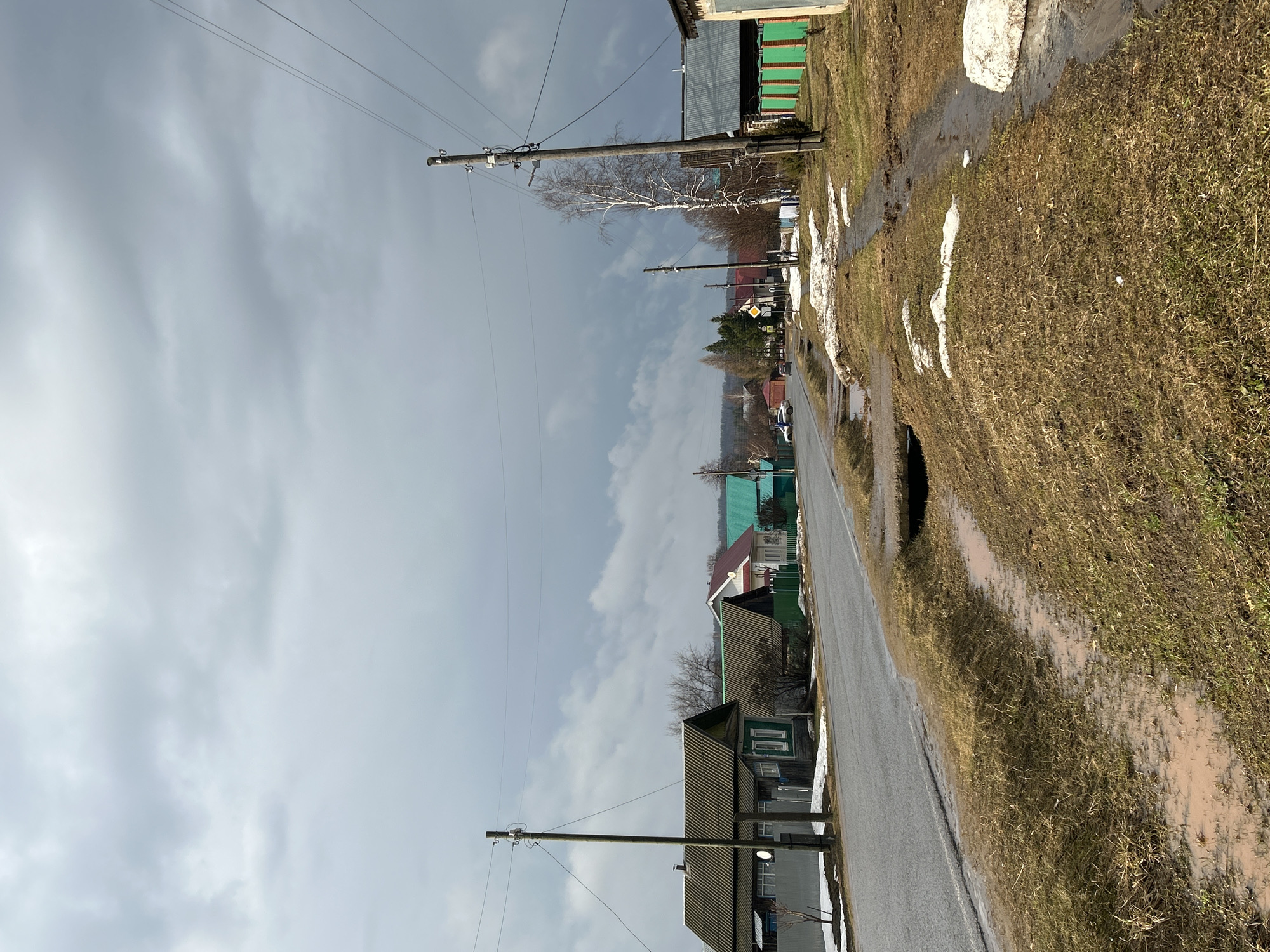 A street in Уральское, Russia. The image shows a rural street with overcast skies. A paved road runs through the center, flanked by grassy areas with patches of melting snow. Wooden houses, some with green trim, line the road. Utility poles with power lines stretch along the street. A road sign is visible further down the road. The vegetation appears sparse and dry, indicative of early spring or late autumn. The ground is wet, suggesting recent rain or snowmelt. No people are visible.