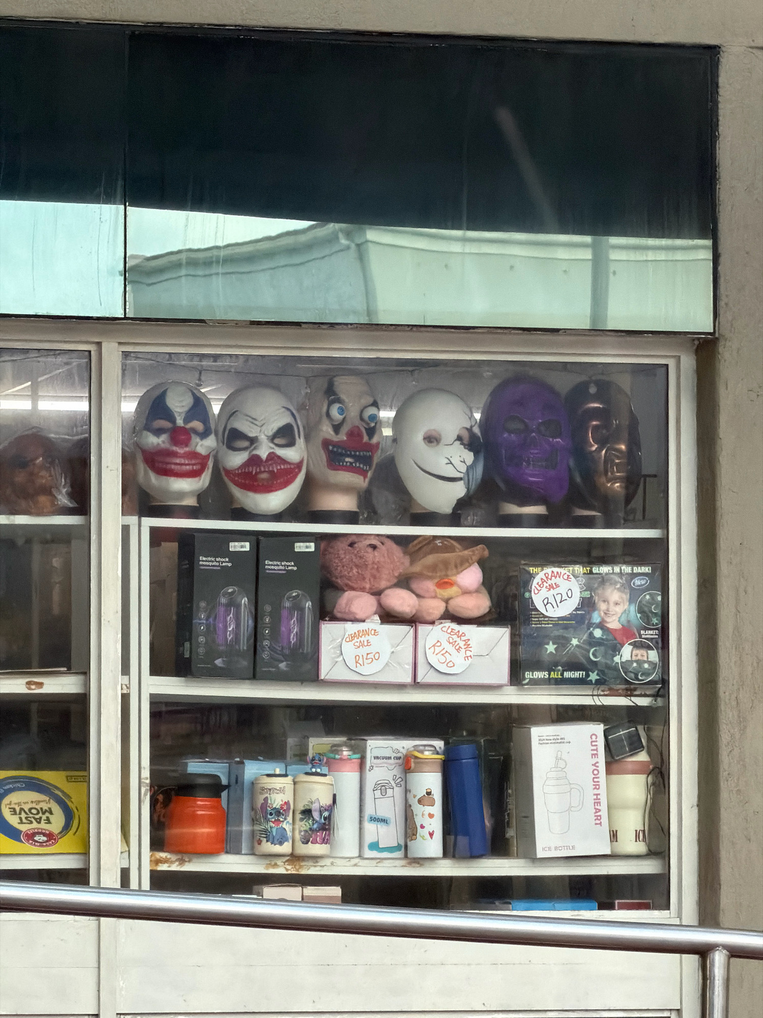 An outdoor retail display cabinet in Fish Hoek, South Africa, showcases a collection of novelty items, primarily masks and novelty drinkware, arranged on shelves. The cabinet has a glass front with metal framing and is divided into two vertical sections.
The top shelf displays several masks, including clown masks with exaggerated features, a white mask with a cracked effect and an unsettling smile, a purple skull mask, and a dark metallic mask. To the left of the masks, a brown, possibly furry, item is partially visible.
The second shelf holds boxes labeled "Electric Shock Mosquito Lamp" with illustrations of the lamps inside. Next to these boxes are two small, stuffed plush toys in a light pink color, along with two white boxes featuring "Clearance Sale R150" labels.
The third shelf is more varied. On the left, a yellow box with blue text and a circular logo that reads "FAST MOVE" is partially visible. Next to it are several drinkware items. A bright orange container is prominent, followed by two cylindrical containers adorned with cartoon characters, possibly Stitch from Disney's Lilo & Stitch. Further to the right are two white vacuum cups, one with blue accents and another with a pink lid. A blue thermos or bottle stands between the cartoon-decorated containers. A box for an "ICE BOTTLE" with the text "CUTE YOUR HEART" is also on this shelf, alongside a beige bottle with a pink lid.
The background visible through the glass includes reflections of the sky, buildings, and possibly a street scene. The overall setting appears to be a storefront or market stall, with the street and pedestrian traffic suggested by the metal railing in the foreground. The lighting is natural, suggesting it is daytime.