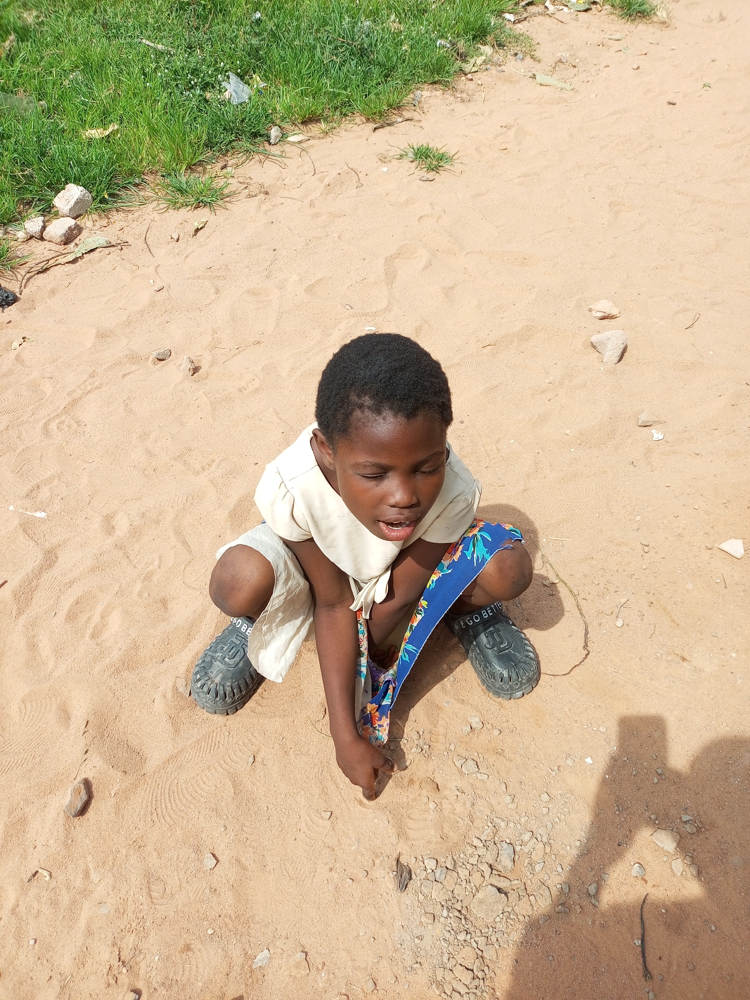 A young child is crouching on sandy ground in Gombe, Nigeria, with a patch of green grass in the background. The child is wearing a light-colored shirt and shorts, with a blue and floral-patterned fabric wrapped around their knees. Dark-colored, open-toed shoes are on their feet, with "GO BETTER" visible on the left shoe. The child's eyes are closed, and their mouth is slightly open. The sandy ground shows footprints and scattered small stones. In the upper left of the image, there are several rocks and a small amount of litter on the grass. The shadow of a person holding a camera or phone is visible in the lower right corner of the frame.