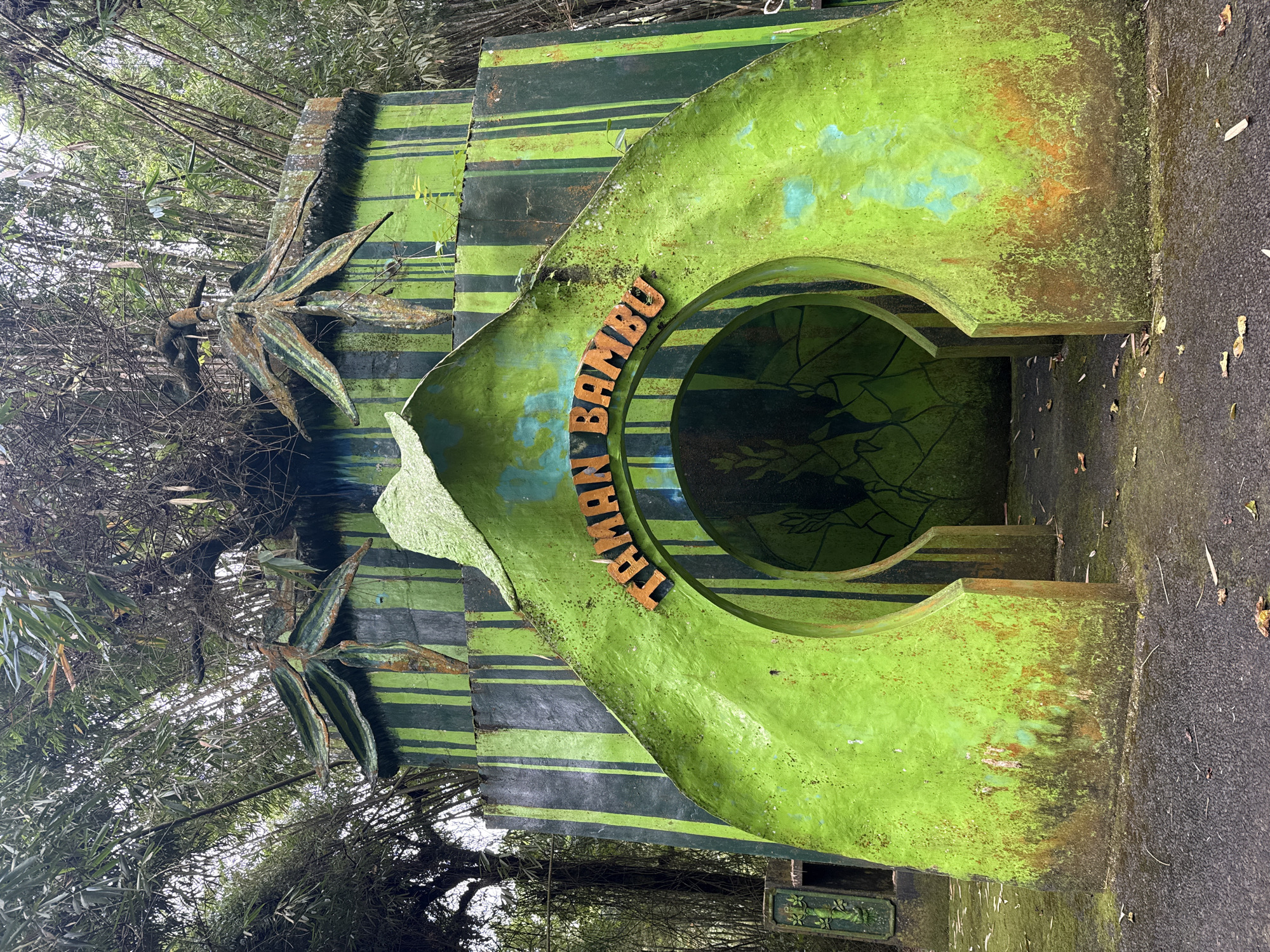 A weathered, green and black sign reading "TAMAN BAMBU" stands in Baturiti, Indonesia. The sign has a circular opening and is surrounded by bamboo trees. The paint is peeling and covered with moss.