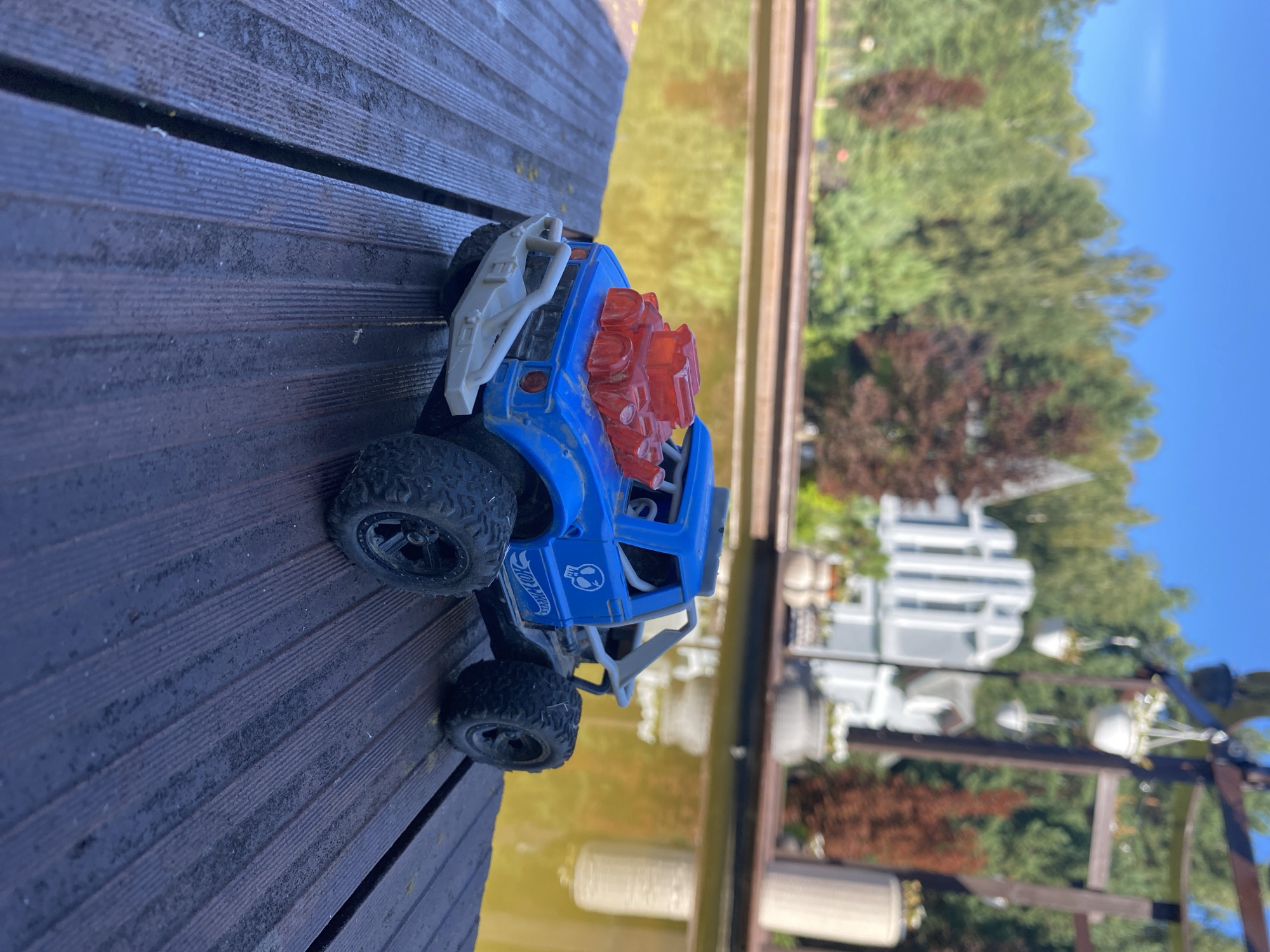A blue toy monster truck with a red engine accessory is positioned on a wooden deck. The truck is facing to the left, with its front bumper visible. Behind the truck, a body of water reflects the surrounding trees and a building. The environment appears to be outdoors, with a clear blue sky overhead. The location is not specified as Chulis, Russia; no geographical context beyond the visual elements is provided. There are no people or interactions depicted in the image.