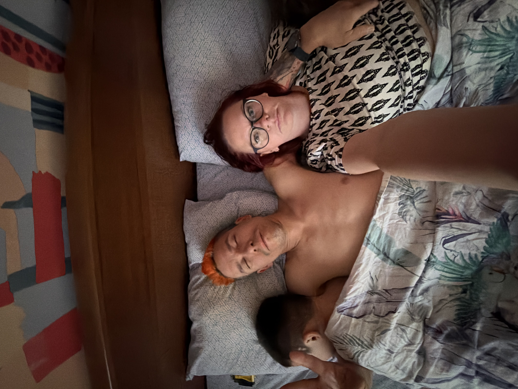 A woman with red hair and glasses, wearing a black and white patterned top, is lying on a pillow in a bed. Beside her, a shirtless man with dyed orange hair is also lying in the bed. A young child is partially visible under the bedsheets, with a hand reaching out. The bed is covered with a white and green patterned duvet. A wall with a geometric design is visible on the left of the frame. This image was taken in Chaikovsky, Russia.