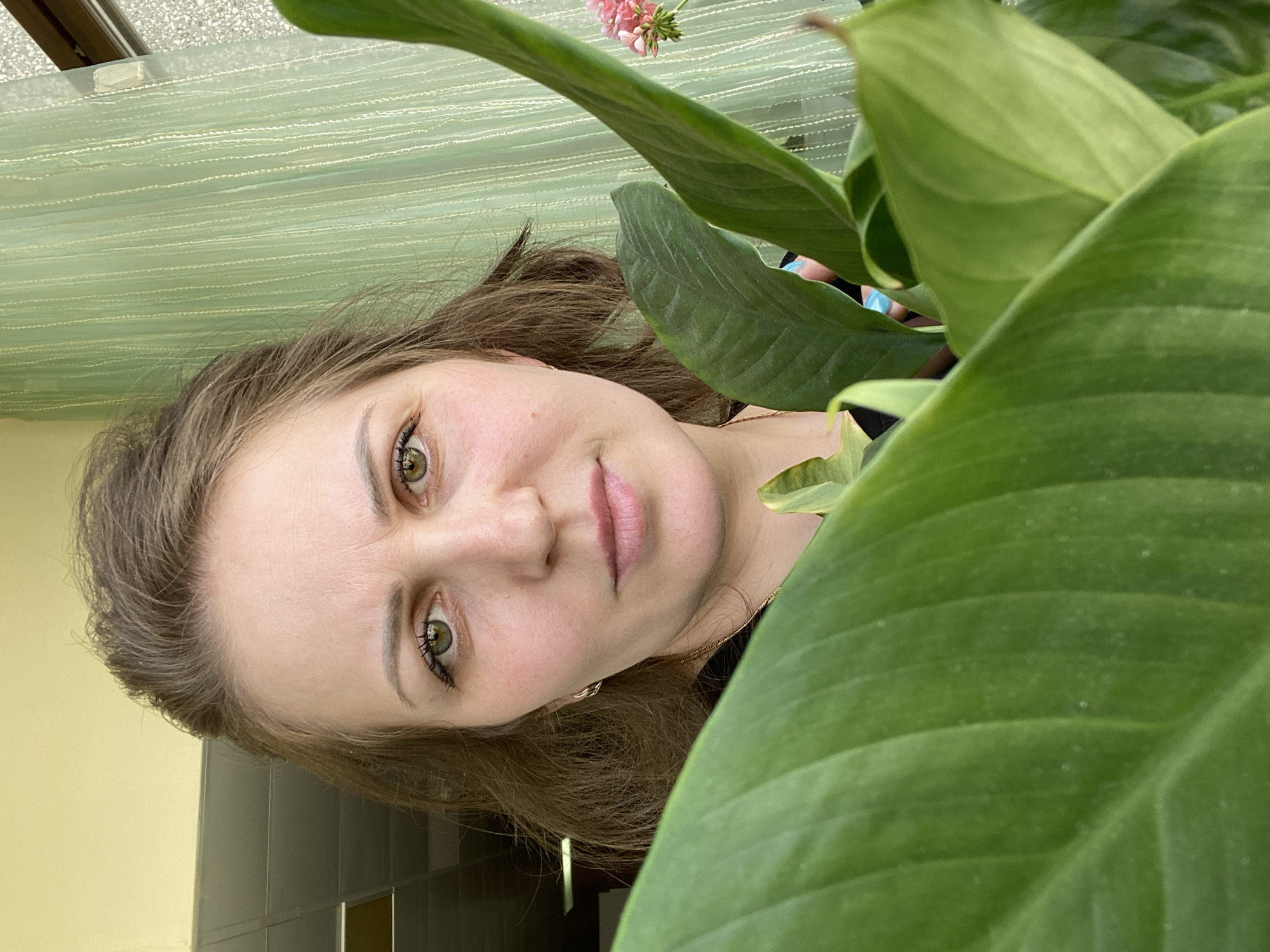 The image displays a headshot portrait of a woman, rotated 90 degrees counter-clockwise. The woman, with light brown hair and green-hazel eyes, looks directly at the camera with a calm, slightly smiling expression. Her face occupies the left and central parts of the frame.
Large, vibrant green leaves of a plant are prominent in the foreground, partially obscuring the right side of the woman's face and the upper right portion of the image. A small cluster of pink flowers is visible behind the leaves in the upper right background.
Behind the woman, a light green, textured curtain or blind with horizontal stripes can be seen, suggesting a window area. A dark window frame is also visible at the top edge of the image. Further in the background, a light yellow wall and a dark furniture piece or partition are present.
The lighting appears soft and natural, indicating an indoor setting during the daytime. The woman seems to be posing for the photo. There is no visible text. Given the context, this scene is situated in Марковский, Russia.