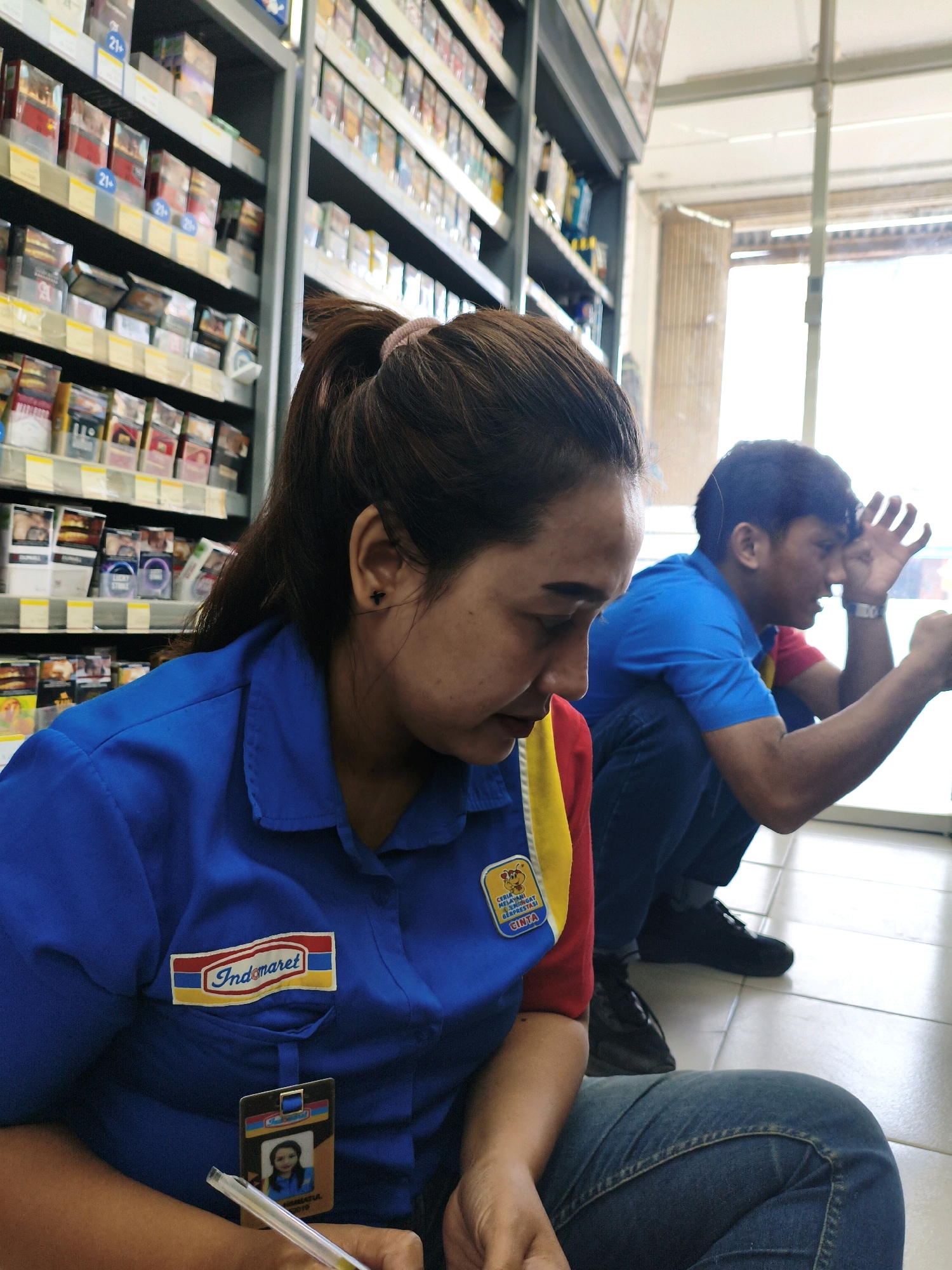 In an indoor convenience store environment, likely located in Sumberejo Kulon, Indonesia, a female employee wearing a blue uniform with "Indomaret" branding and a red and yellow accent on the sleeve is shown from the chest up. She has her hair in a ponytail and is looking down, appearing to be filling out paperwork with a pen. A lanyard with an ID card, featuring her photo, is attached to her uniform. Behind her and to her right, a male individual in a blue shirt with a red and yellow collar is crouched down, looking towards his right with his hand raised near his forehead. The background shows shelves stocked with various products, including what appear to be cigarette packs and other retail items. The flooring is tiled. The lighting suggests it is daytime, with natural light coming from a doorway or window in the background.
