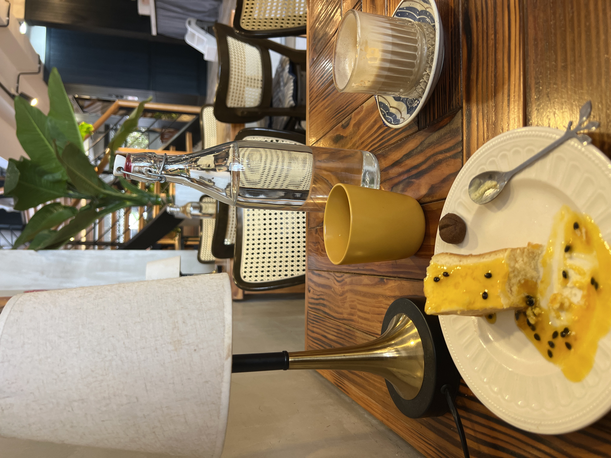 An eye-level shot captures a cozy indoor cafe or restaurant setting in Bắc Nha Trang, Vietnam, featuring a wooden table laden with refreshments. The rich, dark brown grain of the tabletop is prominently visible.
In the foreground, a white fabric lampshade with a striking brass-colored, horn-shaped base provides ambient light, its black electrical cord trailing. Next to the lamp, a white ceramic plate holds a slice of light-colored cake, generously covered with vibrant yellow passion fruit pulp and dotted with black seeds. A small, dark brown cookie or truffle accompanies the cake, and a silver spoon with a delicate leaf-shaped handle rests on the plate.
Further back on the table, a clear glass bottle with a swing-top closure, filled with water, stands alongside a mustard-yellow ceramic mug. A ribbed clear glass containing a milky beverage, possibly coffee or tea, is perched on a blue and white patterned saucer with intricate designs.
The background of the scene features a blur of the interior, including several chairs with dark wooden frames and woven rattan or cane backs, suggesting comfortable seating for patrons. Lush green foliage from a large houseplant, possibly a bird of paradise, adds a natural element to the warm indoor ambiance. The overall impression is one of leisure and relaxation, inviting a moment of enjoyment with dessert and drinks. No visible text is present.