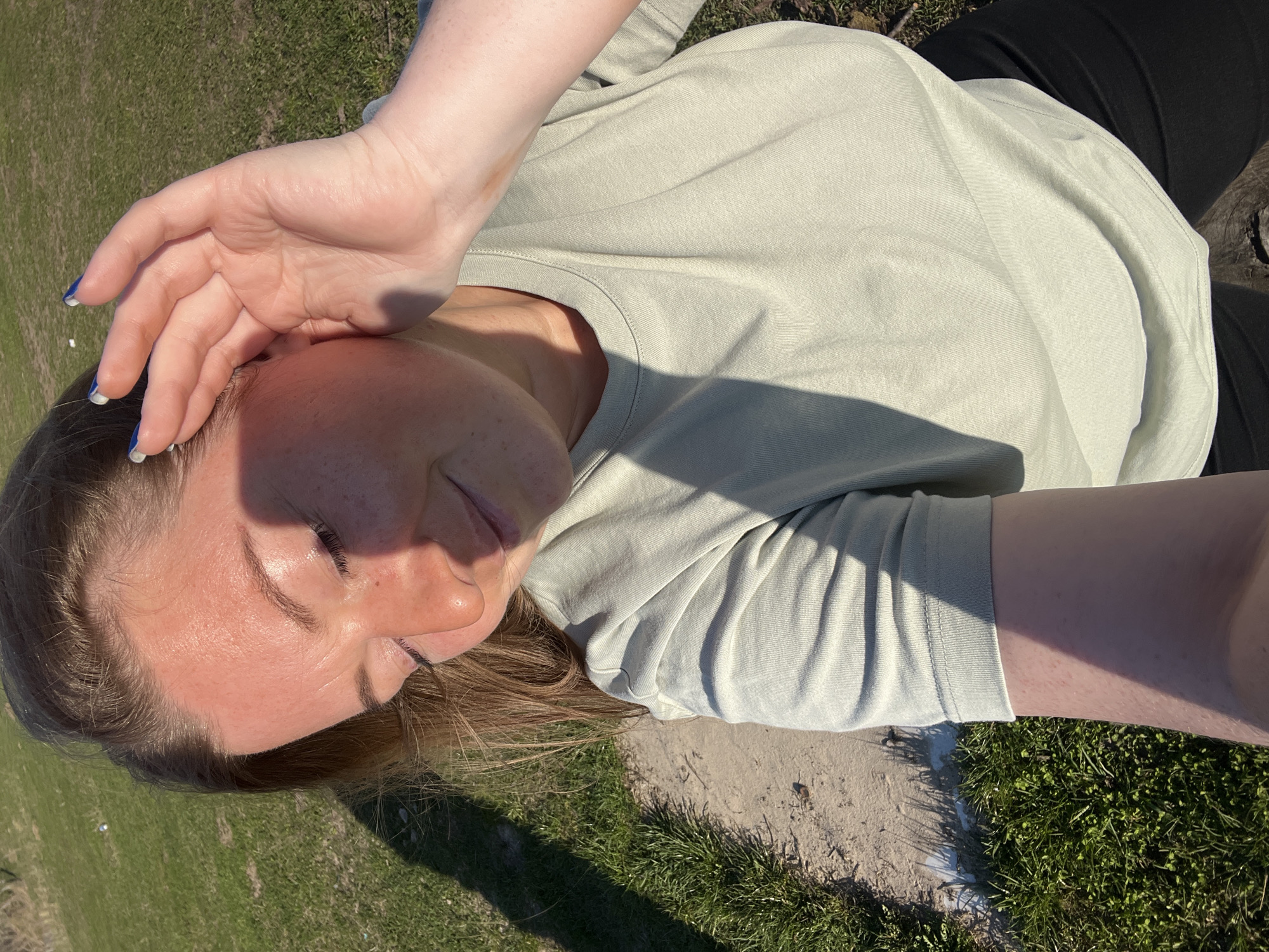 A woman is lying on the grass in Раздолье, Russia. She is wearing a light green long-sleeved shirt and black pants. Her left hand is positioned over her forehead, shielding her eyes from the sun. The location appears to be a park or grassy area near a concrete path. The image is taken from a slightly elevated, angled perspective.
