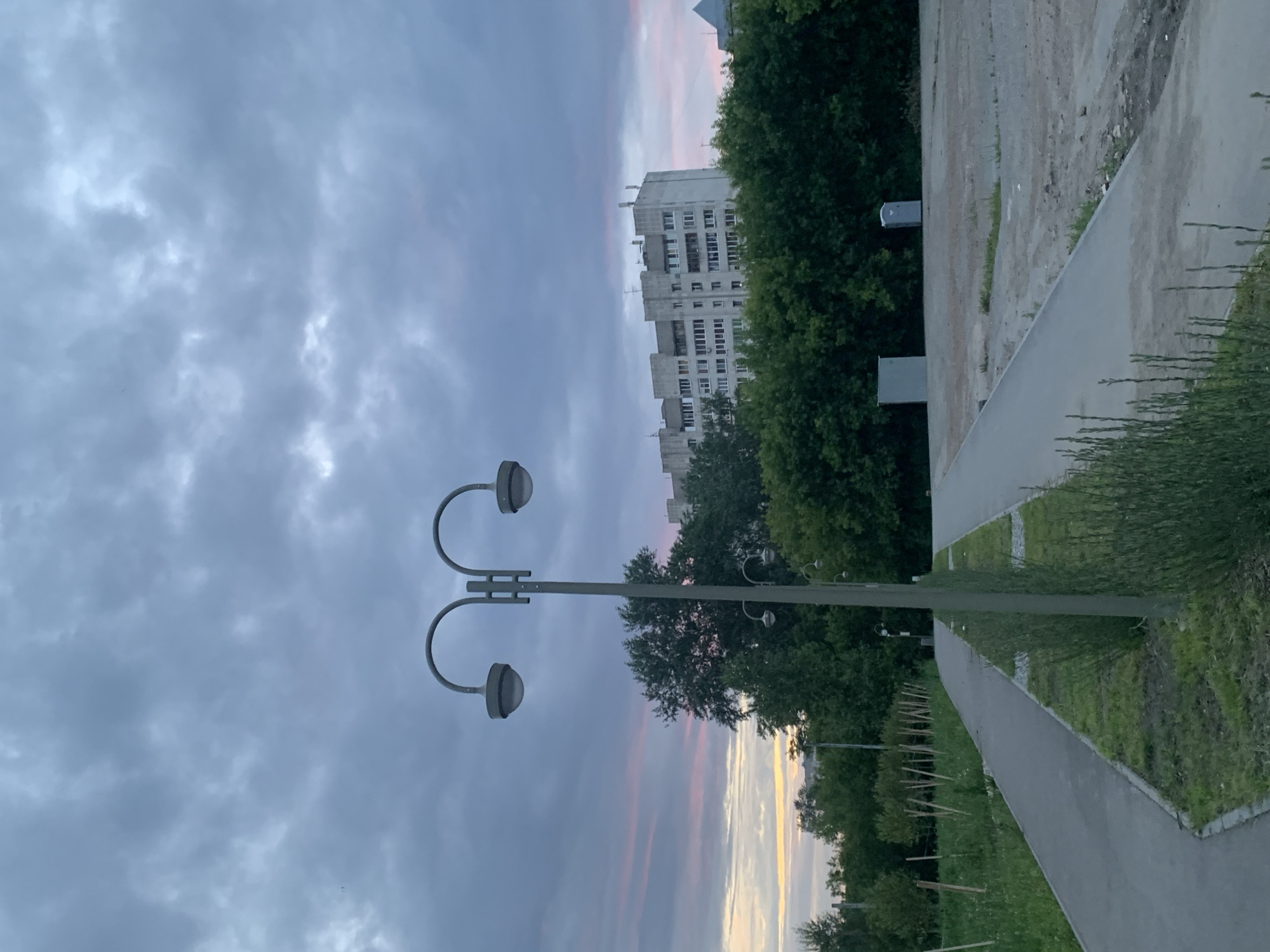 A dual-headed street lamp stands prominently in the foreground, its gray metal pole rising vertically. The sky above is overcast with dark gray clouds, tinged with faint pink and yellow hues near the horizon, suggesting a twilight or dawn setting. Behind the street lamp, a dense line of green trees forms a backdrop. Beyond the trees, a multi-story, light-colored apartment building with numerous windows is visible. In the middle ground, a paved pathway curves from the left towards the right, bordered by grass and low-lying vegetation. To the right of the pathway, a large, open area of dirt and gravel is visible, with two portable structures, likely toilets, situated near the tree line. The overall scene appears to be an outdoor public space in an urban or suburban environment. The location is identified as the Perm State Technical University campus, specifically along the western pathway of the campus, near the student dormitory complex.