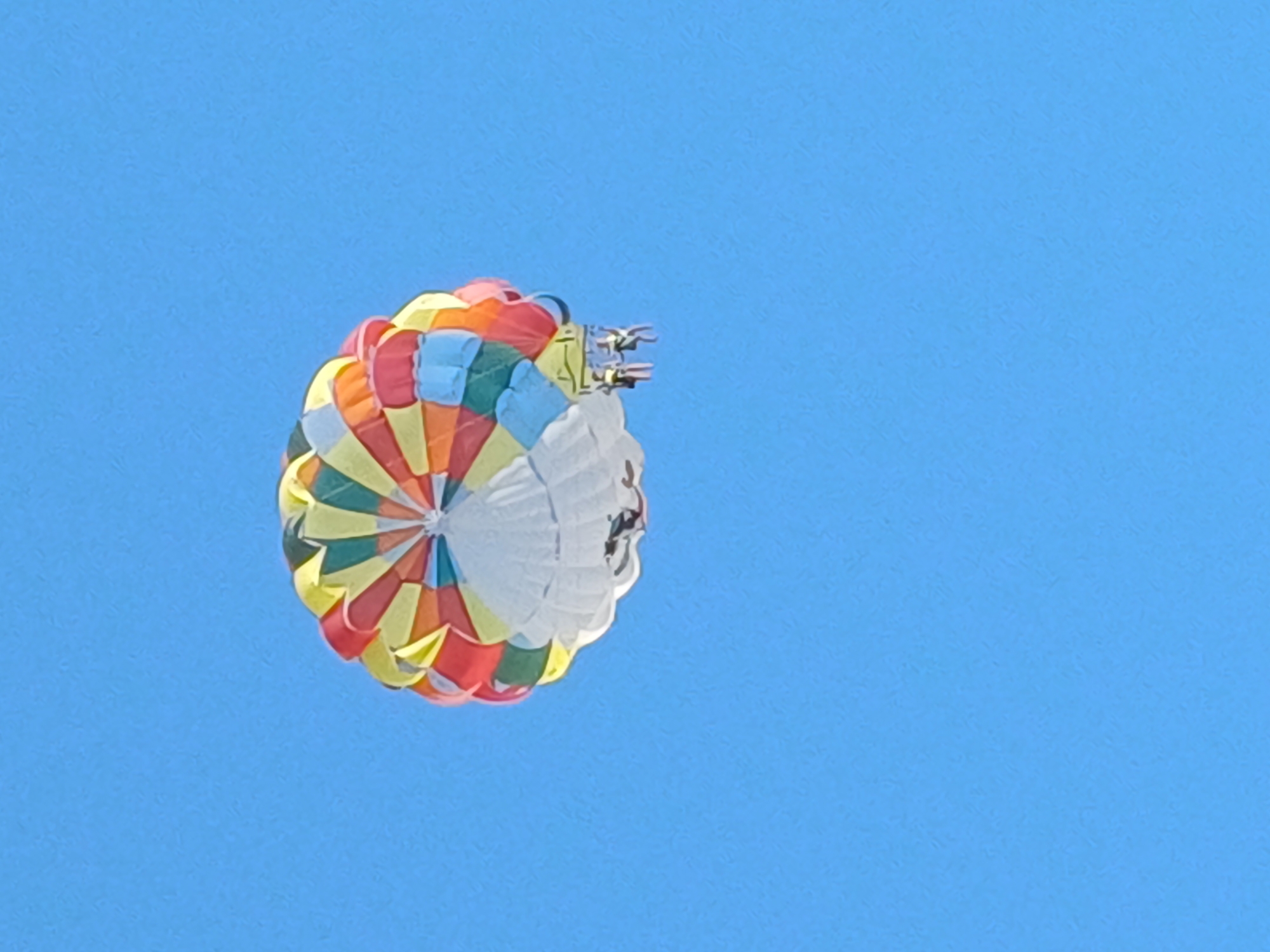 The image shows a colorful parasail flying in a clear blue sky. The parasail is a large parachute-like wing with segments of various bright colors, including red, orange, yellow, green, and blue, arranged in a circular pattern. At the top of the parasail, a small harness is visible with two people appearing to be strapped in and enjoying the ride. The scene suggests a recreational activity, likely taking place on a sunny day. There are no visible text elements in the image. The setting is a vast, uninterrupted expanse of blue sky, offering no specific geographical cues other than the general impression of an open, outdoor environment suitable for parasailing. The activity is parasailing, where individuals are towed behind a boat while suspended from a parachute-like wing. The overall impression is one of leisure and adventure in good weather conditions.