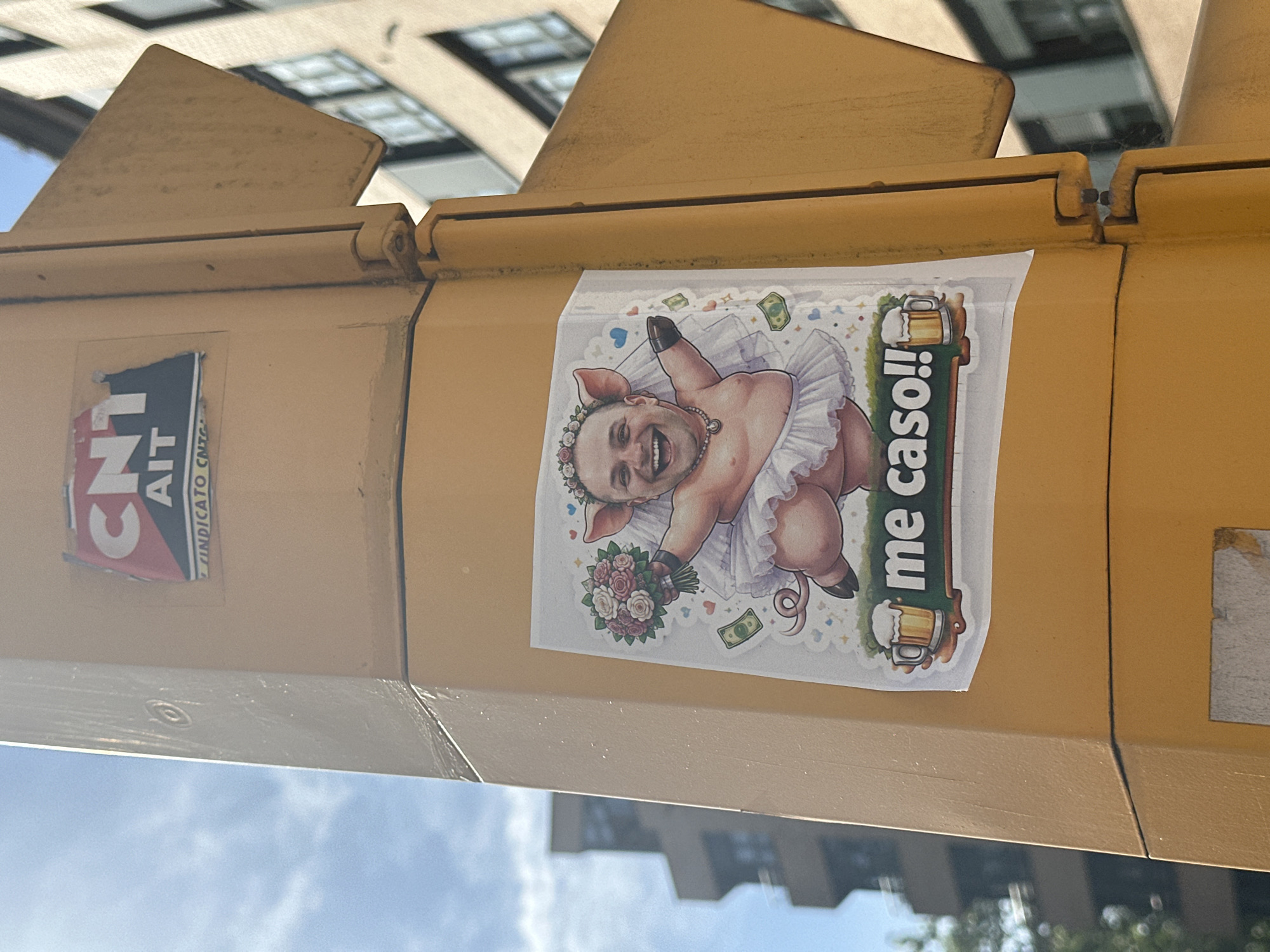 The image shows a yellow metal traffic light post in Gijón/Xixón, Spain. A CNT-AIT sticker is attached to the left. On the right, a sticker depicts a pig with a human face, wearing a tutu and a flower crown, holding a bouquet, with the text "me caso!!" and images of beer mugs and money. Buildings and sky are visible in the background.