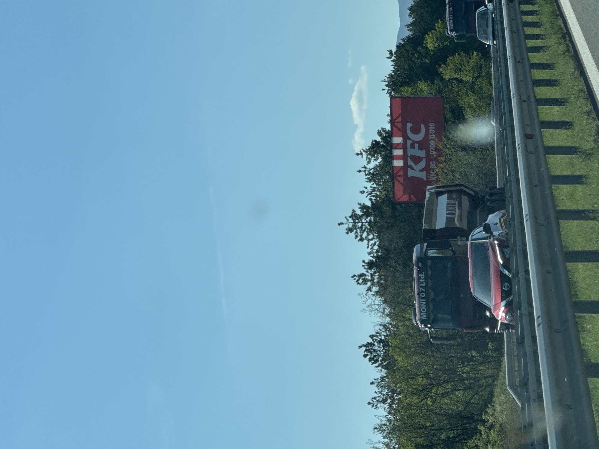 The image depicts a daytime scene on a multi-lane highway, observed from a vehicle. The sky is clear and bright blue with a single small white cloud.
To the right of the highway, a large red billboard is prominently displayed, featuring the "KFC" logo in white. Below the logo, smaller white text reads "KFC.BG" and a phone number, "8709 11 999". A white glare or lens flare is visible on the billboard's surface.
Vehicles are traveling on the highway. A large dark truck, with "MONI 07 Ltd." visible on its side, is in the foreground. Behind the truck, a red SUV, possibly a Nissan model due to its headlight design, is also present. Other vehicles are visible further down the road, indicating traffic.
Along the right side of the highway, dense green trees and vegetation line the route, with mountains or hills visible in the distant background. A metal guardrail separates the highway lanes from the roadside. The setting appears to be a sunny day in Lesichovo, Bulgaria, consistent with the "KFC.BG" on the billboard.