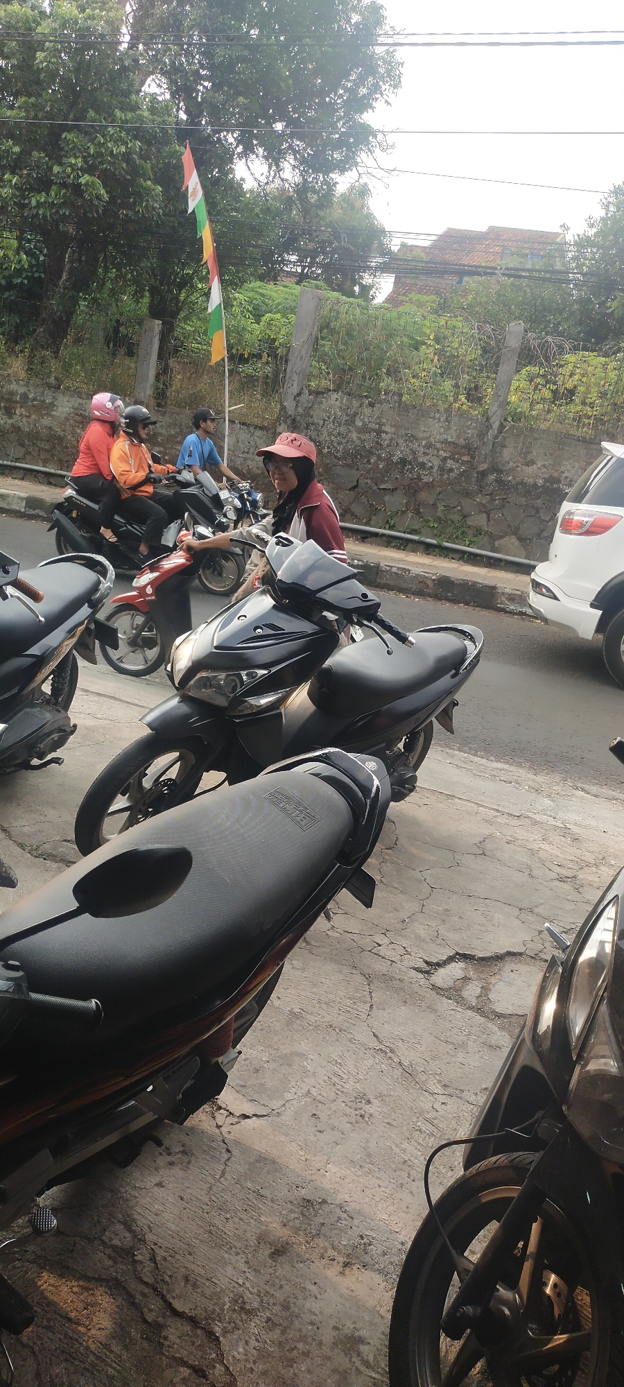In Cimaung, Indonesia, on a paved street with a stone retaining wall and some greenery, several motorcycles are parked and in motion. A white SUV is visible on the right side of the street. In the foreground, a black motorcycle's seat and front wheel are prominent. Further back, a rider in a red cap and jacket is on a motorcycle, looking towards the camera. To their left, three individuals are on two motorcycles, two of whom are wearing helmets. A flag with multiple colored stripes is flying from a pole on the left side of the frame. The sky is bright and overcast.
