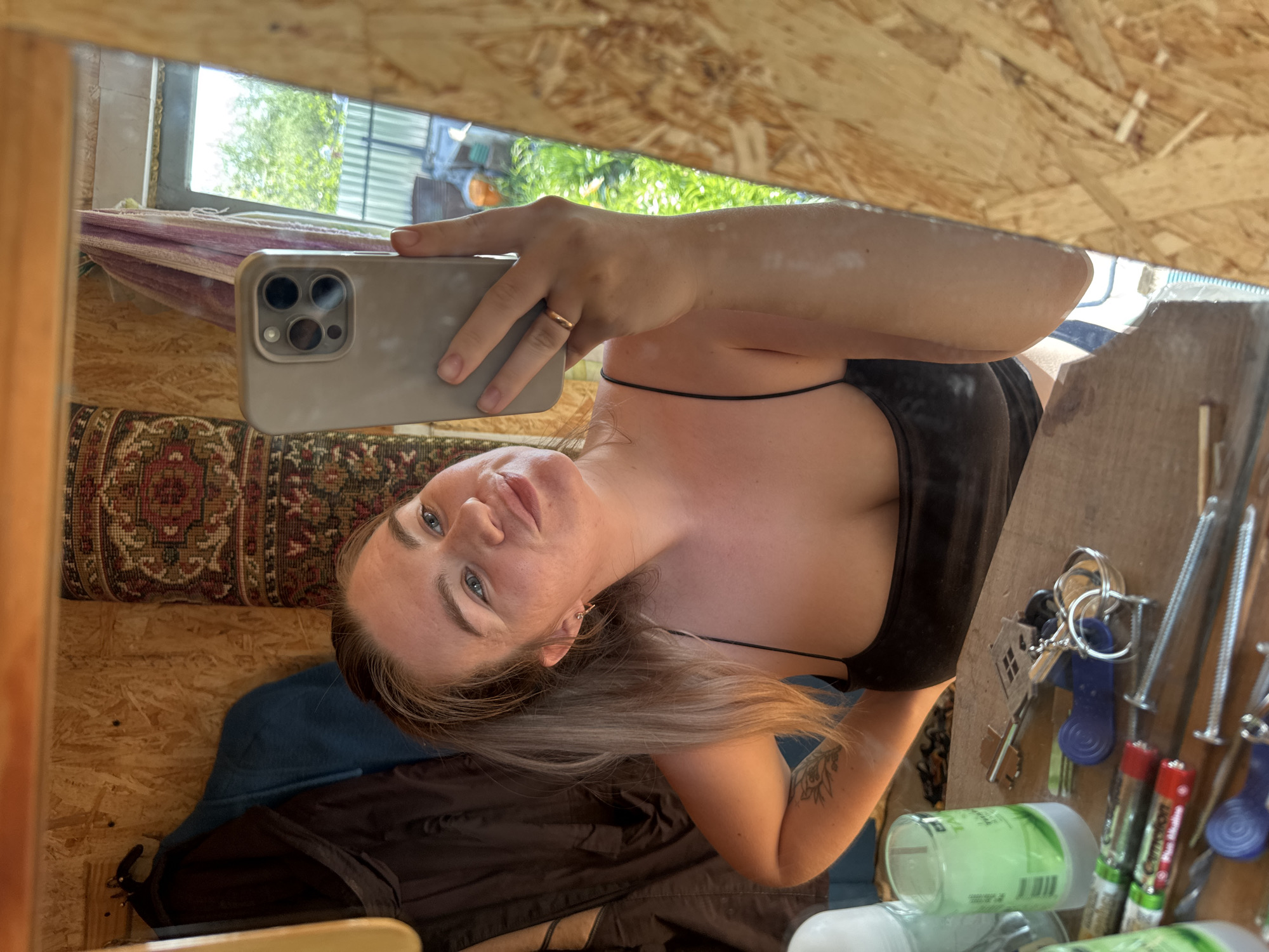The image shows a woman in a black tank top taking a selfie in a mirror. She is holding a smartphone with a triple-lens camera. The background includes a wooden wall, a window showing greenery outside, and a cluttered shelf with keys, tools, and various items. The setting appears to be a shed or workshop. The location is in the city of Chaikovsky, Russia.