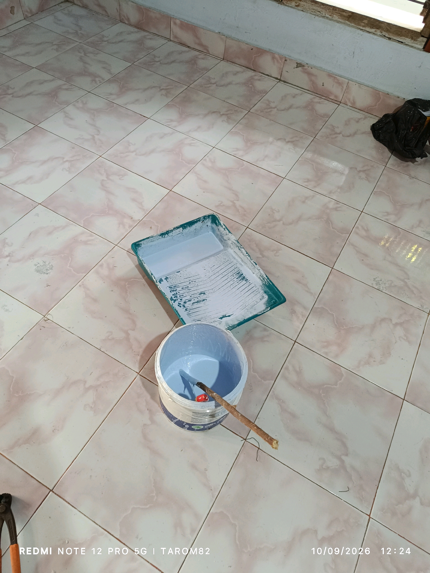The image depicts a floor with light-colored tiles featuring pinkish marbling. In the center of the frame, there is a green paint tray with white paint residue and a white bucket containing light blue paint. A wooden stick, possibly a stirrer or a paint roller handle, rests across the rim of the bucket. To the upper right, a black plastic bag is partially visible. In the lower left corner, the top portion of a pair of pliers with orange handles can be seen. The bottom of the image displays a timestamp and device information: "REDMI NOTE 12 PRO 5G | TAROM82" and "10/09/2026 12:24". The environment appears to be indoors, possibly during a painting or renovation project, with a tiled floor and a wall with a window ledge visible in the background. The image was taken in Rejoagung, Indonesia.