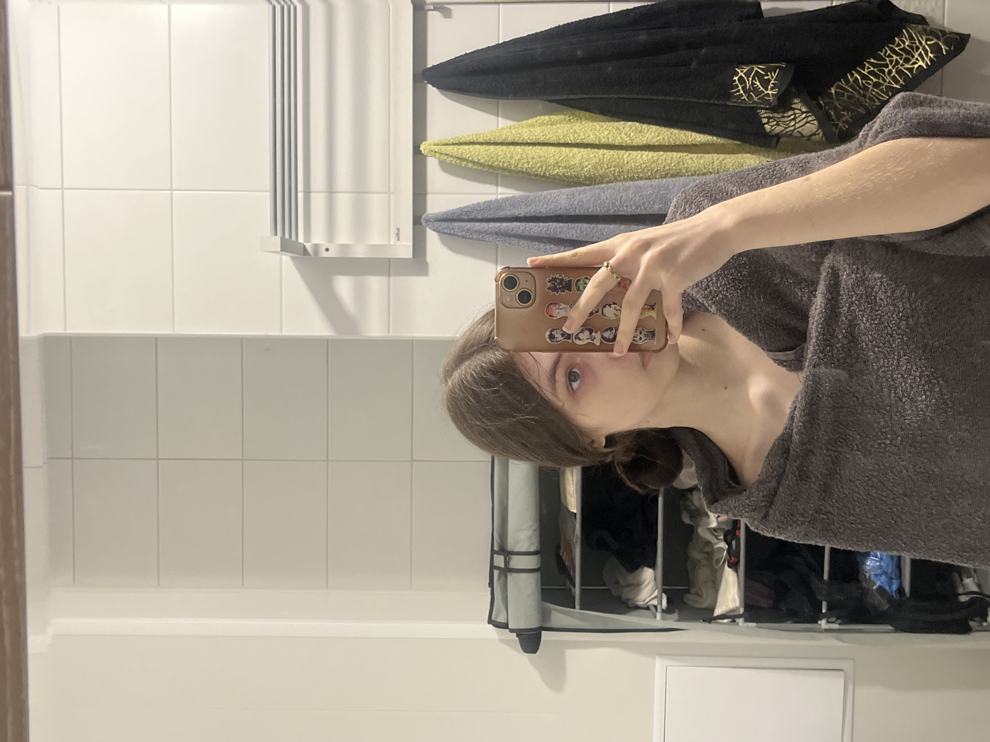A mirror selfie captures a young person, likely female, in a bathroom. The person has light brown hair and is seen from the chest up, wearing a grey terry cloth robe or towel wrap. They are holding a golden-brown smartphone to take the picture, with part of their face, including one eye, visible in the reflection. The phone case is decorated with multiple small, colorful stickers depicting various anime or cartoon characters.
The background features white tiled walls with a white baseboard running along the bottom. To the left, a white panel, possibly a towel heater or radiator, is mounted on the wall. Above and to the right of the person's reflection, three towels are neatly folded and hanging. The top towel is black with an intricate golden pattern, followed by a light greenish-yellow towel, and then a plain grey towel. Below these towels, an open grey shelving unit holds various folded items, possibly clothes or laundry. A white electrical outlet or cover is visible on the lower right portion of the wall. The lighting appears to be artificial indoor light. The image is taken in Saint Petersburg, Russia.