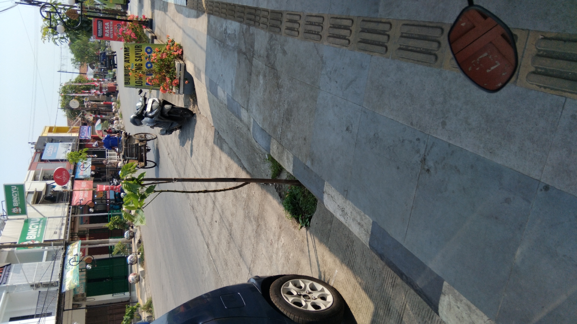 A street scene in Batang, Indonesia, shows a paved road with several businesses lining the left side. In the foreground, a dark blue car is parked on the left, its rear wheel visible. A young tree stands on the sidewalk to the right of the car, with its shadow falling across the road. Further down the street, a man in a blue shirt pushes a bicycle with a cart attached. Several motorcycles are parked along the sidewalk, and a vendor stall selling flowers and Indonesian signage is visible on the right. The sky is clear and blue, indicating a sunny day. The environment suggests a commercial area with pedestrian and vehicle traffic.