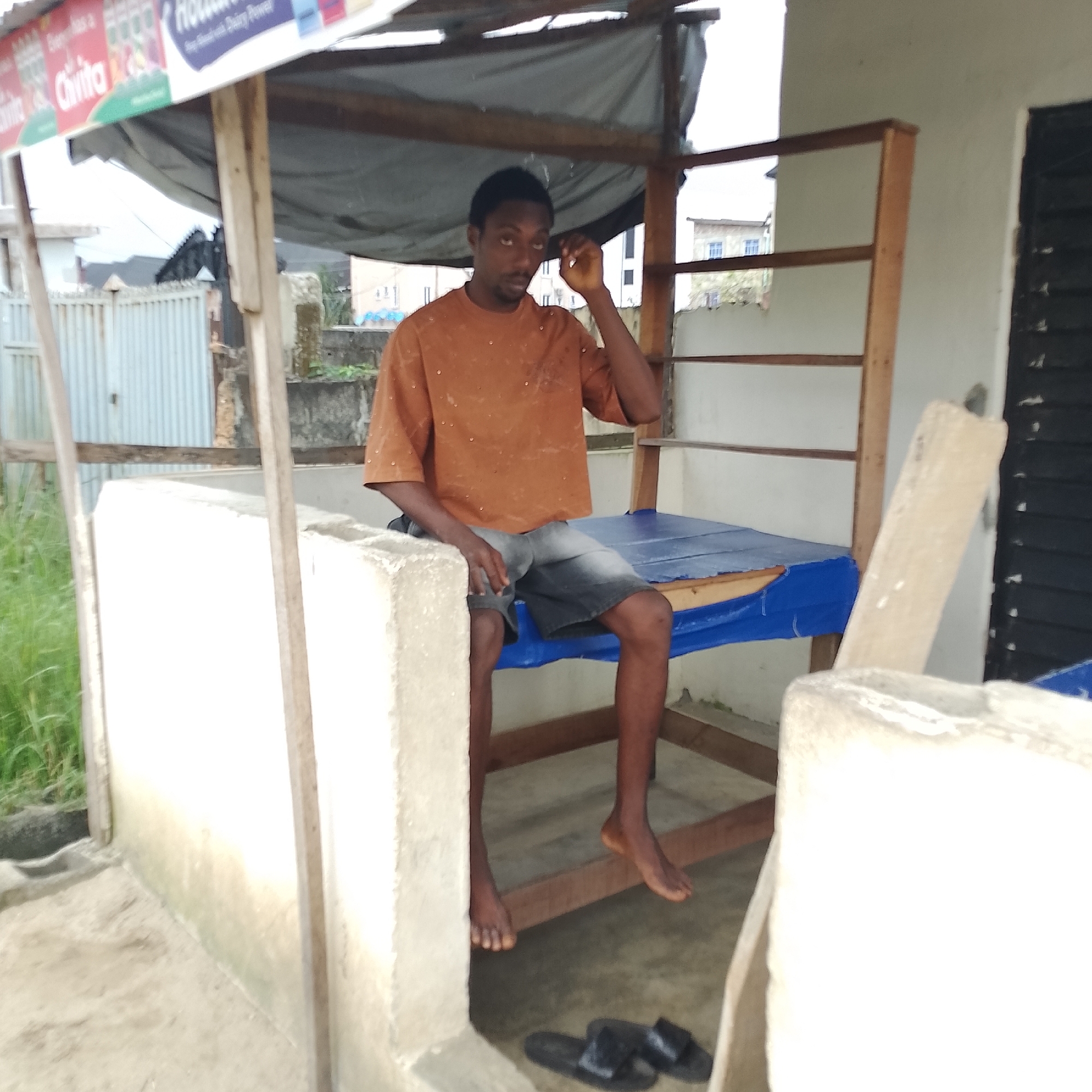 In Ado, Nigeria, a young man is seated on a blue tarp-covered structure, likely a bed or bench, inside a makeshift stall. He is wearing an orange t-shirt with small embellishments and dark denim shorts. The stall has a corrugated metal roof supported by wooden beams and a concrete wall on the left. In the background, a corrugated metal fence and buildings are visible. A pair of black sandals rests on the ground near the man's feet. The overall environment appears to be a humble outdoor setting.