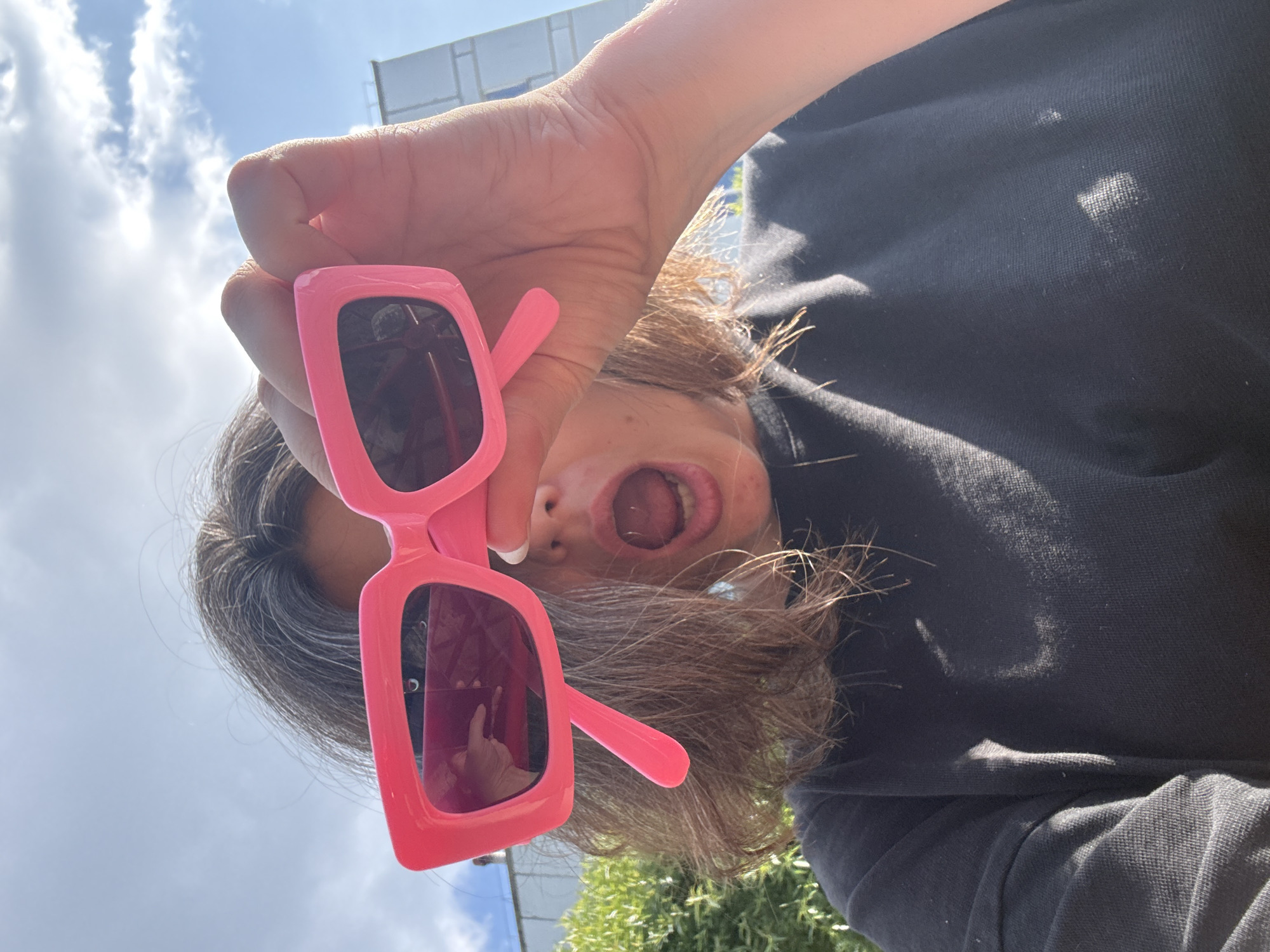 A low-angle, close-up shot captures a person's face from the chest up, outdoors on a sunny day. The person, with brown hair and visible grey streaks, is holding a pair of bright pink, rectangular sunglasses up to their face, obscuring their eyes. Their mouth is open wide in an expression of surprise or excitement. They are wearing a dark crewneck sweatshirt. The background shows a bright blue sky with scattered white clouds, some greenery on the left, and a white building with blue trim on the right. The scene is likely a public outdoor space in Chaikovsky, Russia, suggested by the presence of residential buildings and vegetation typical of urban or suburban areas.
