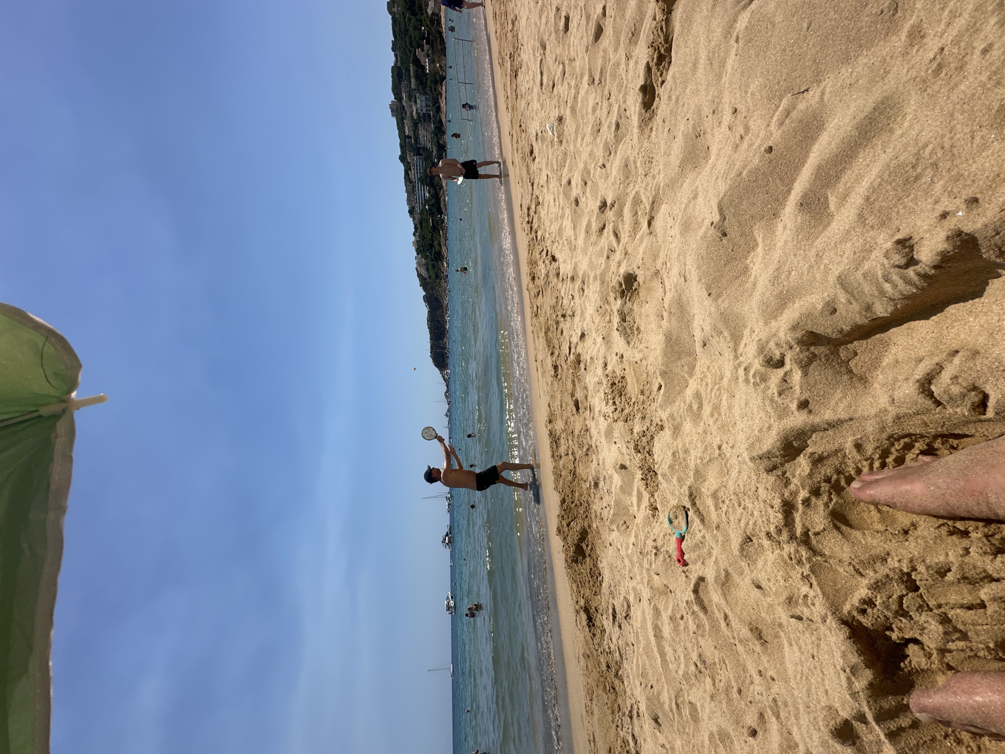 A daytime, eye-level shot shows a sandy beach bordering a body of water. Two young men are playing paddle ball on the wet sand where the water meets the beach. One man, on the left, is wearing dark shorts and a dark cap, and is in mid-action, swinging a paddle. The other man, on the right, is also wearing dark shorts and is standing, holding a paddle. Several other people are visible in the water behind them, some swimming and others on boats. The water is calm and blue, and beyond that, a hillside with buildings and vegetation can be seen. The foreground of the image is dominated by sand dunes, with a child's red and blue plastic spade lying on the sand. The sky is clear and blue. The overall scene depicts a lively beach day, likely in Sant Feliu de Guíxols, Spain, judging by the Mediterranean coastline visible.