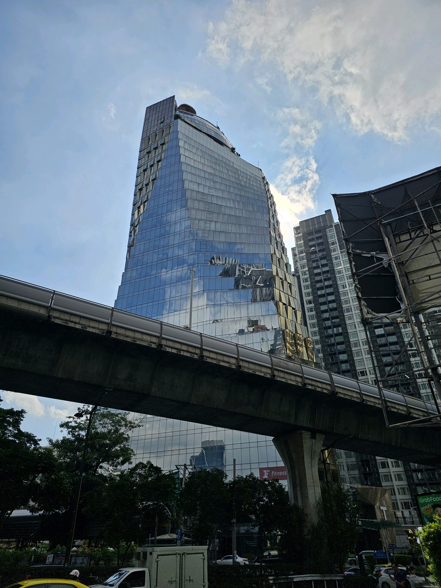 The image depicts a modern cityscape in Bangkok, Thailand, characterized by towering skyscrapers and elevated infrastructure. The central focus is a striking, angular glass building with a distinctive twisted design, reflecting the sky and surrounding structures. To the right of this building, another, taller skyscraper with a grid-like facade of windows is visible. In the foreground, an elevated train track structure, supported by concrete pillars, runs horizontally across the frame, partially obscuring the lower portions of the buildings. The sky is a mix of blue with scattered white clouds. Green trees line the street level, and several vehicles, including a white truck and a yellow car, are present on the road below. A sign with the word "Fitness" is visible on the glass building. The overall scene suggests a busy urban environment.