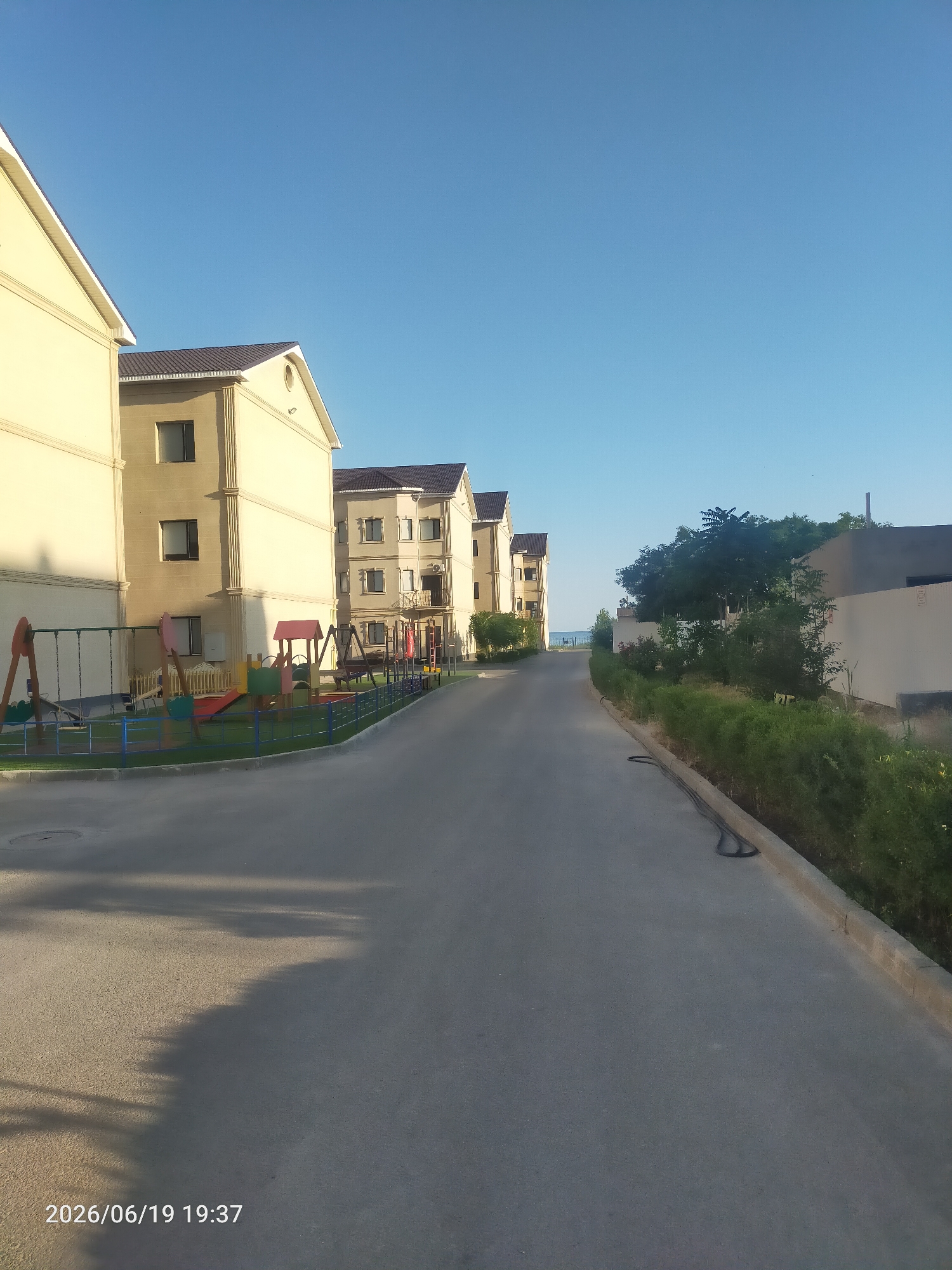 The image depicts a residential street lined with pale yellow apartment buildings. In the foreground, a children's playground with swings, a slide, and climbing structures is visible. The buildings are modern in design with dark tiled roofs and regularly spaced windows. The street is paved and appears to be well-maintained. To the right of the street, there is a dense line of green bushes and trees, and beyond that, a wall of a different building. In the distance, the sky is a clear, bright blue, suggesting fair weather. The lighting indicates it is late afternoon or early evening, with long shadows cast across the street. The overall impression is one of a peaceful and tidy residential area. There is visible text in the bottom left corner of the image that reads "2026/06/19 19:37".