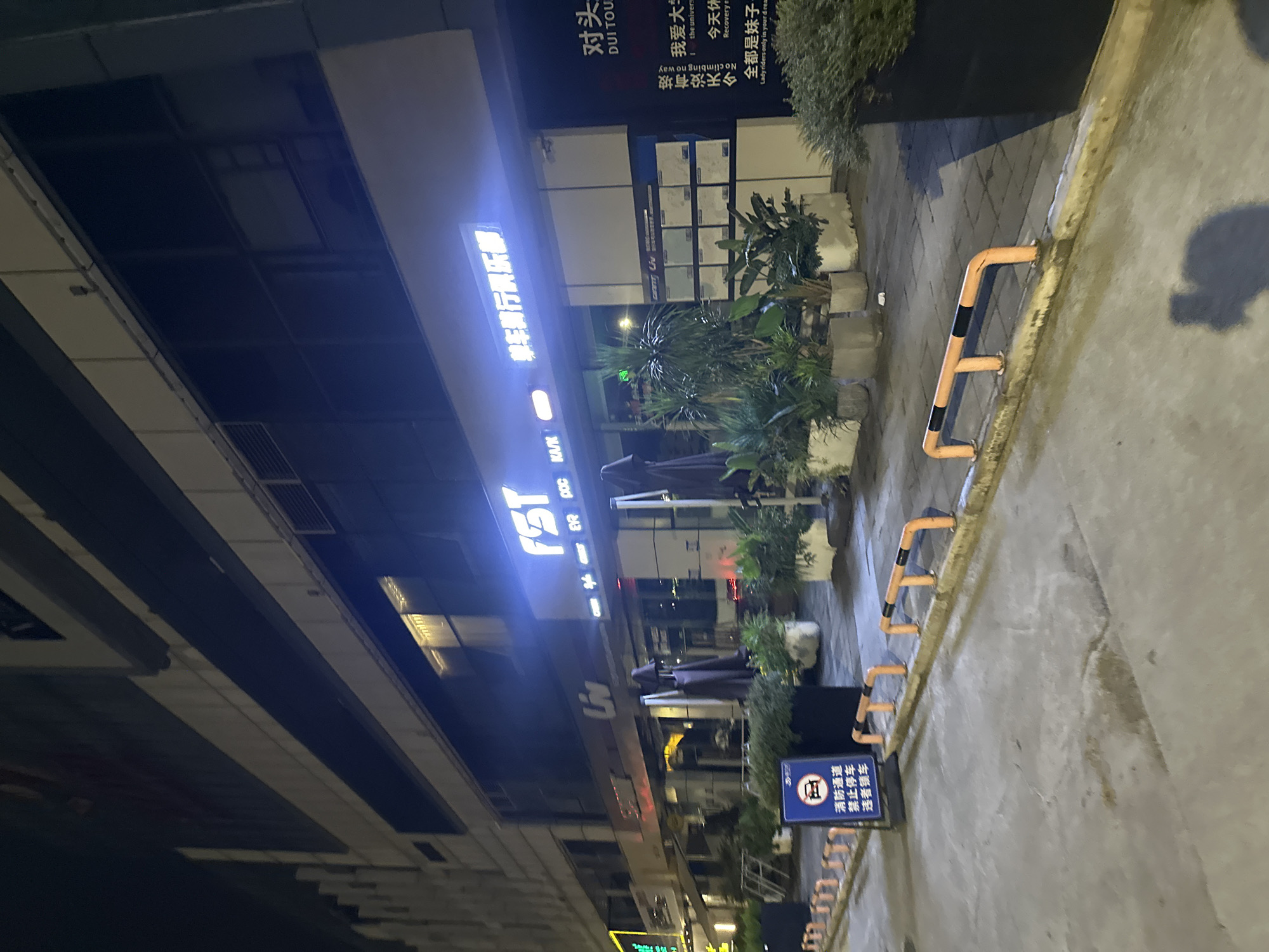 This image captures a nighttime street scene in the Chongqing High-tech Zone, China, featuring the exterior of a business establishment. The building's facade is illuminated by neon signage and interior lighting, reflecting on the wet pavement. A prominent illuminated sign reads "FST" with accompanying Chinese characters. Various potted plants are strategically placed along the sidewalk, with dark umbrellas leaning against the building. In the foreground, a blue sign with Chinese text and a car icon indicates parking restrictions, likely related to fire safety, prohibiting parking in the area. Orange and black barriers line the edge of the sidewalk. The environment appears to be an urban commercial district.