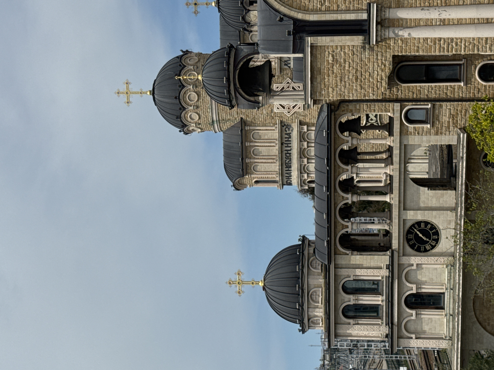 The image shows the Cathedral of Michael the Archangel in Sochi, Russia. The cathedral features dark domes topped with golden crosses, a clock tower with a round clock face, and brick and stone walls with arched windows and ornate detailing. The sky is light blue. The image is vertically oriented.