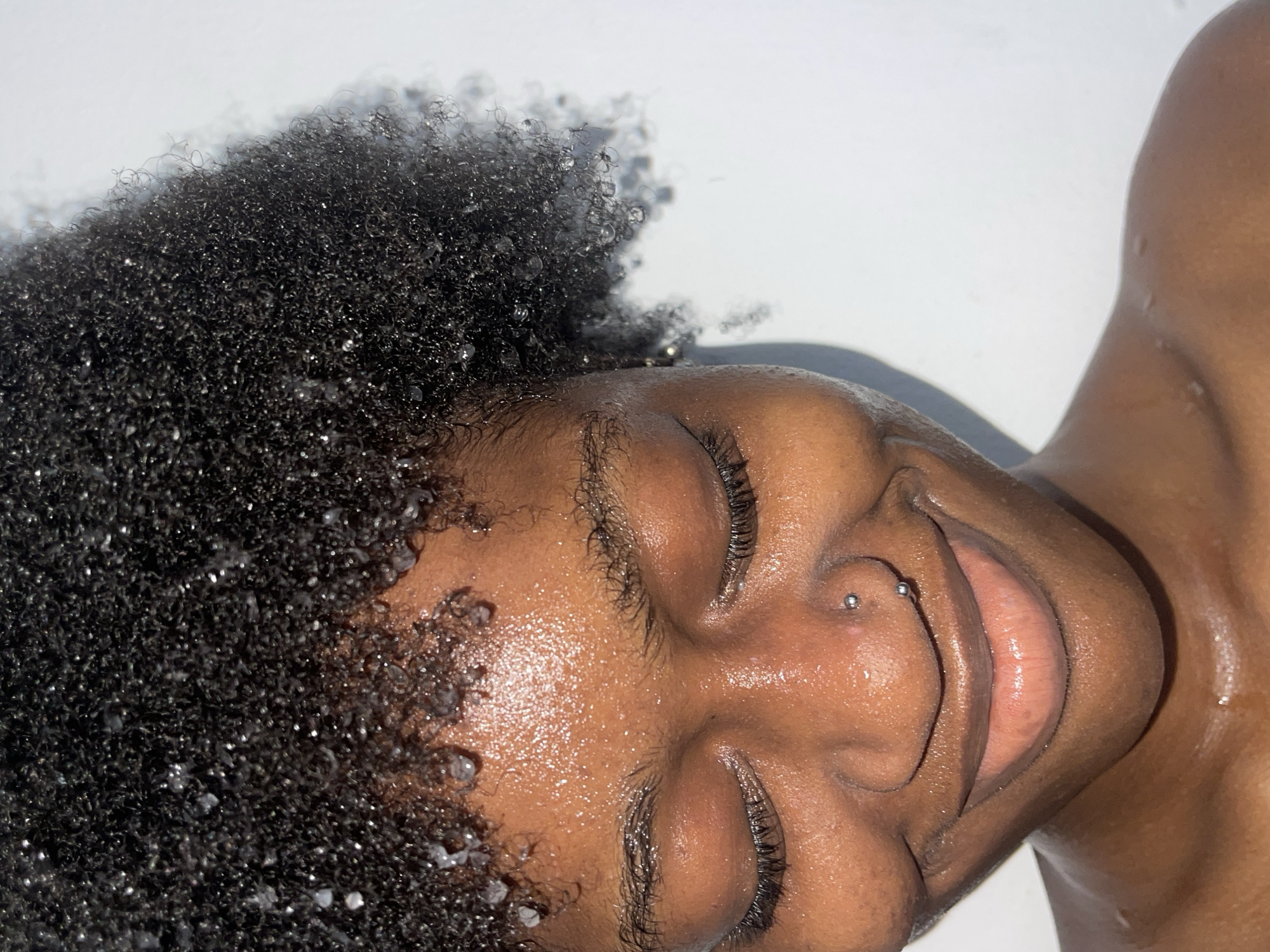 A close-up shot captures an individual with dark, afro-textured hair wet with droplets, lying on a white surface. The person's eyes are closed and they are smiling, with a nostril piercing visible. The skin appears moist and reflective. Part of the shoulder is visible. This scene is not discernible as being in Ado, Nigeria.