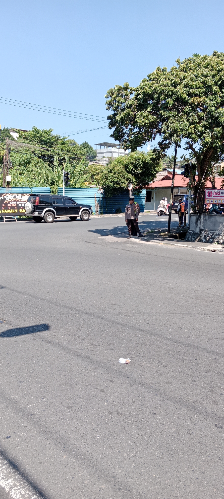 The image depicts an outdoor scene on a clear, sunny day in Tanjung Pinang, Indonesia. The foreground is dominated by an asphalt road, with a light gray surface and some scattered debris. The middle ground features an intersection with a black SUV parked on the left side of the road. Two individuals in police uniforms are visible; one stands near the SUV, and another is further to the right, near a traffic light pole. Behind them, there is a teal-colored fence or barrier. Beyond the barrier, buildings with red roofs and green foliage from trees are present. A tall, white, multi-story structure with a visible roof deck is seen in the background, partially obscured by trees. The sky is a clear, bright blue. A traffic signal is visible on a pole near the intersection. A motorcycle is also present in the mid-ground, to the right of the teal barrier, with a rider. Another person in an orange vest is standing to the right of the traffic light pole.