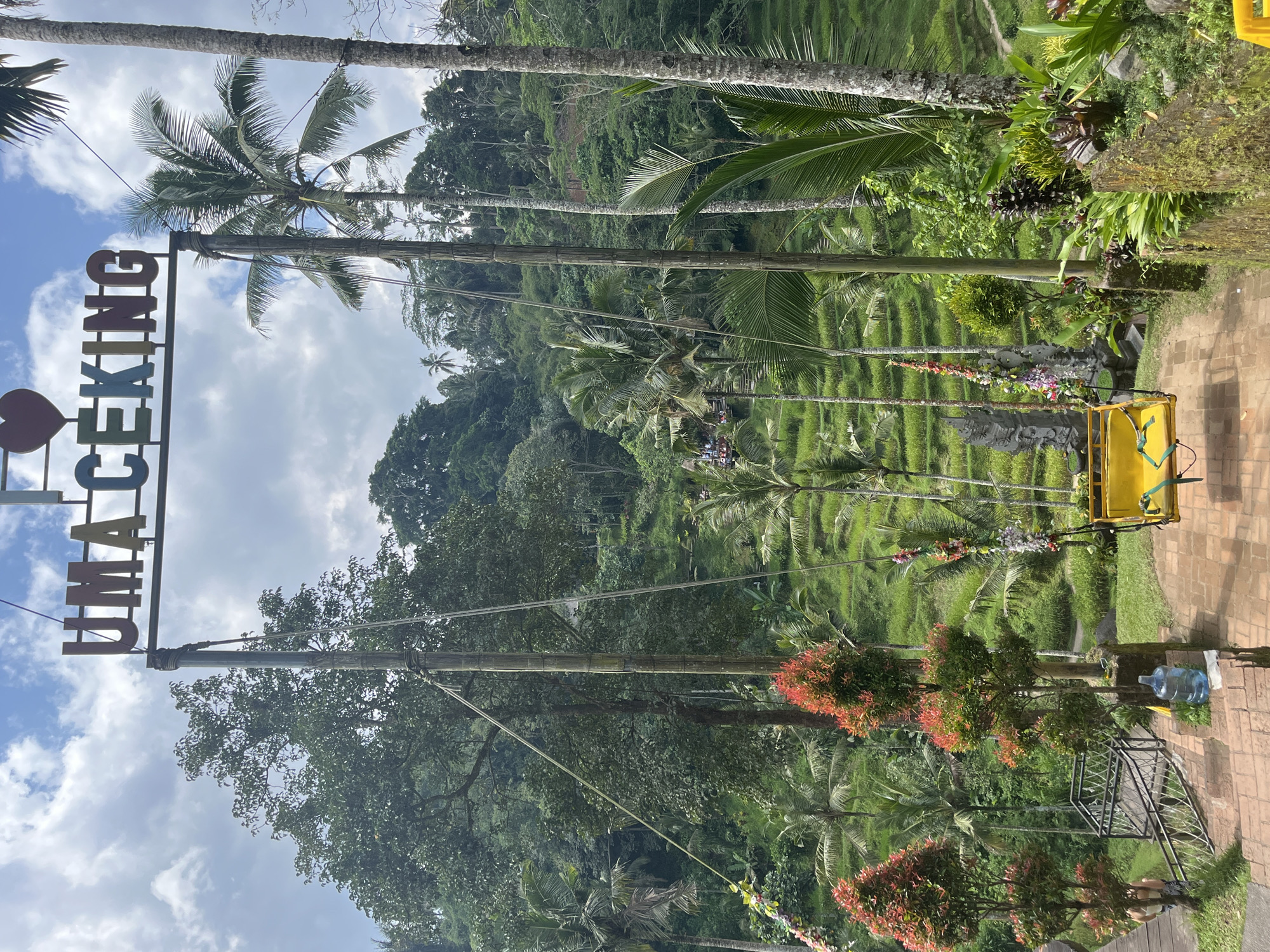 The photograph shows a view of the Tegallalang Rice Terraces in Tegallalang, Indonesia. In the foreground is a sign reading "UMA CEKING" in vertical letters, with a heart above the word "UMA." A tall bamboo structure holds up the sign and a large yellow swing. The swing is adorned with flowers. The background features terraced rice paddies, lush green vegetation, and palm trees. The sky is partly cloudy with blue patches. A brick path is visible on the right side of the frame. A water bottle is on the ground.