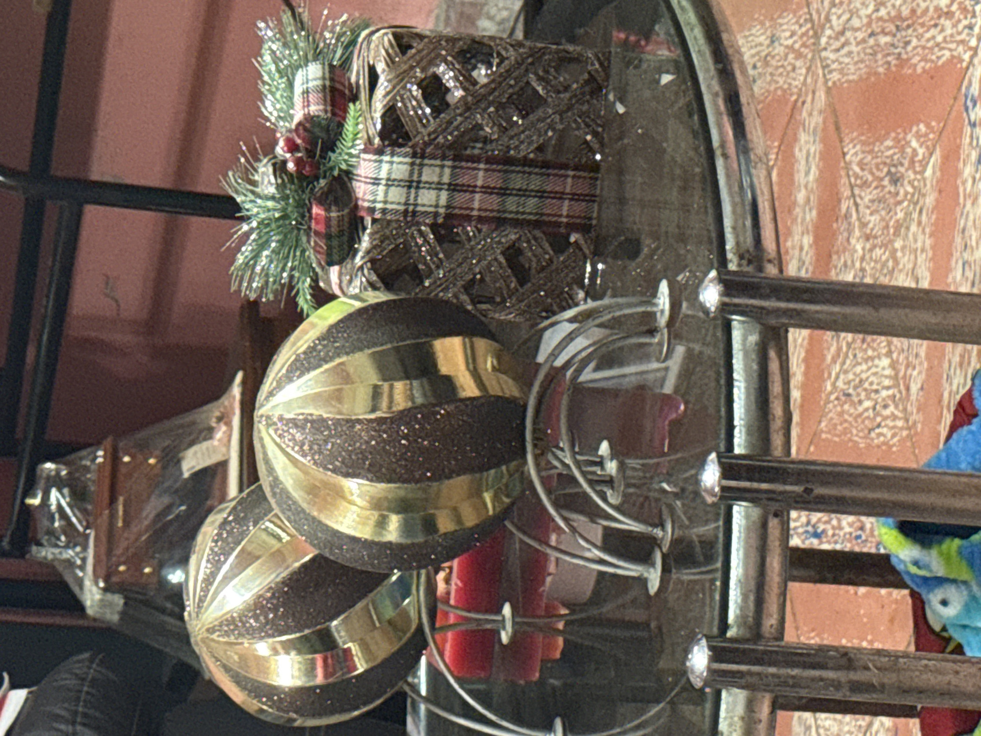 The image shows a still life on a glass-topped metal table. Two brown and gold Christmas ornaments rest on the table, near some red candles. A Christmas decorative item with greenery and a plaid ribbon is positioned near the center of the table. A wrapped wooden box is partly visible behind the ornaments. The background features a pinkish-red wall and other furniture. There is no indication as to the image's connection to San Cristóbal, Mexico. No people are visible.
