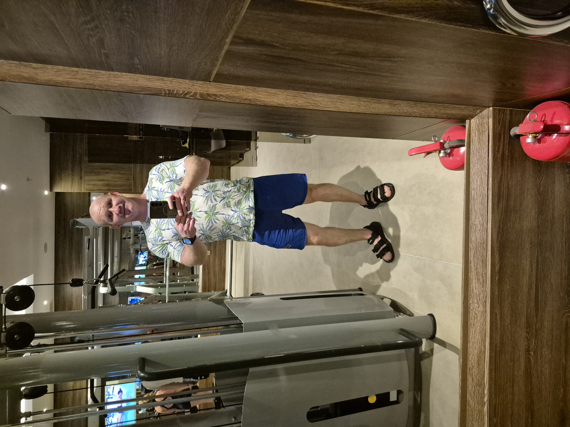 The image shows a man in a gym taking a mirror selfie. He is wearing a short-sleeved floral shirt, blue shorts, and black sandals. The gym is equipped with weight machines and televisions. There are two red fire extinguishers mounted near the mirror.
The location is likely in Alanya, Turkey, given the user's request.