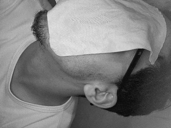 A person lies down with a white cloth covering their face and head. Dark hair and beard are visible. Glasses frame the side of their face, resting behind the ear. The person wears a white tank top, revealing the neck and shoulder. The image is in black and white, highlighting textures and contrasts. The focus is on the head and upper body, emphasizing the resting posture.