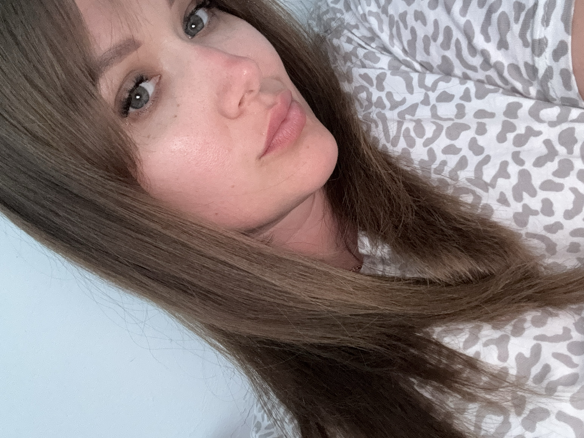 A close-up portrait of a woman with shoulder-length brown hair and bangs. She has fair skin, grey eyes, and full lips. She is wearing a white top with a grey leopard print. The background appears to be a light blue wall. The location is unknown.