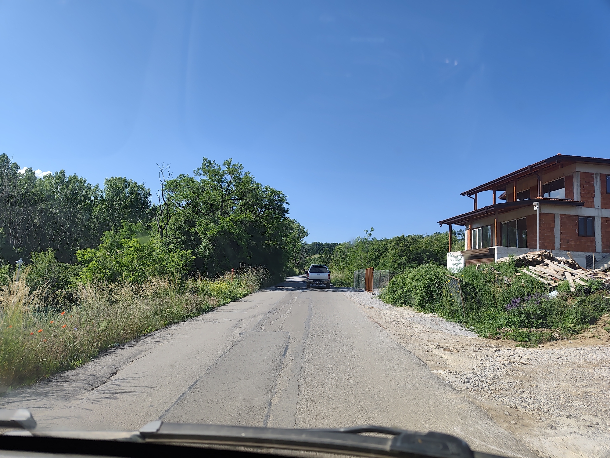 The image depicts a rural road in Rudartsi, Bulgaria, on a clear, sunny day. The asphalt road is in moderate condition, showing some cracks and patches. On the left side of the road, tall, dry grass and wildflowers, including red poppies, line the verge, with dense green trees forming a backdrop. On the right side, a partially constructed brick building with wooden roof beams and a balcony is visible. A pile of lumber is stacked near the building. The building is surrounded by green vegetation and gravel. A white SUV is driving away from the camera down the road. The scene appears to be captured from the perspective of a vehicle, with the windshield wipers visible in the foreground. The sky is a clear, bright blue.