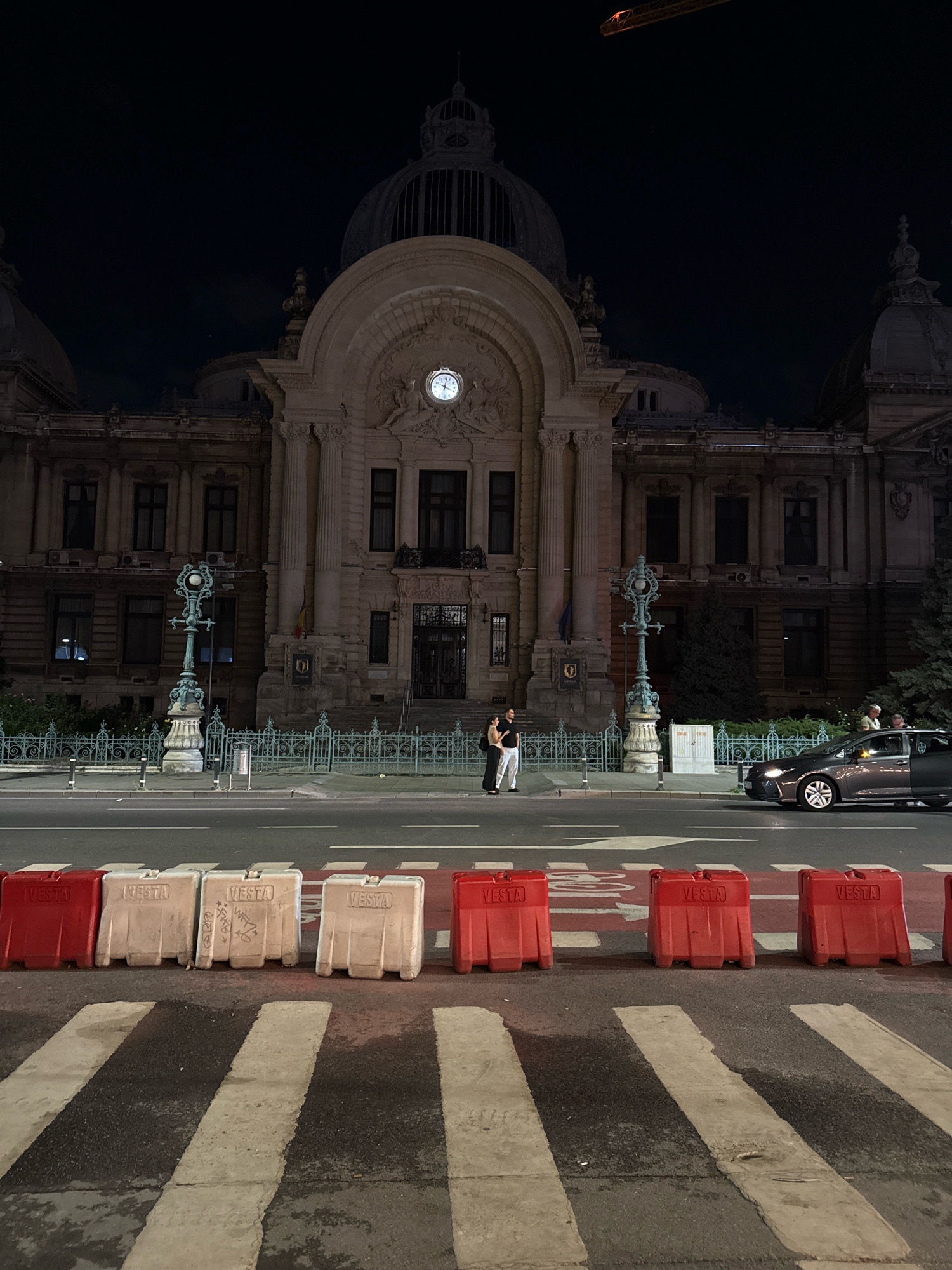 The image shows the Bucharest National Bank building at night, a grand edifice with classical architecture. A large clock is visible on its facade. In the foreground, a street is marked with a pedestrian crossing, bordered by red and white plastic barriers. Two people are standing near the building's entrance, embracing. A car is driving on the road. The scene is lit by streetlights and the illumination from the building itself, creating a nighttime urban atmosphere.
