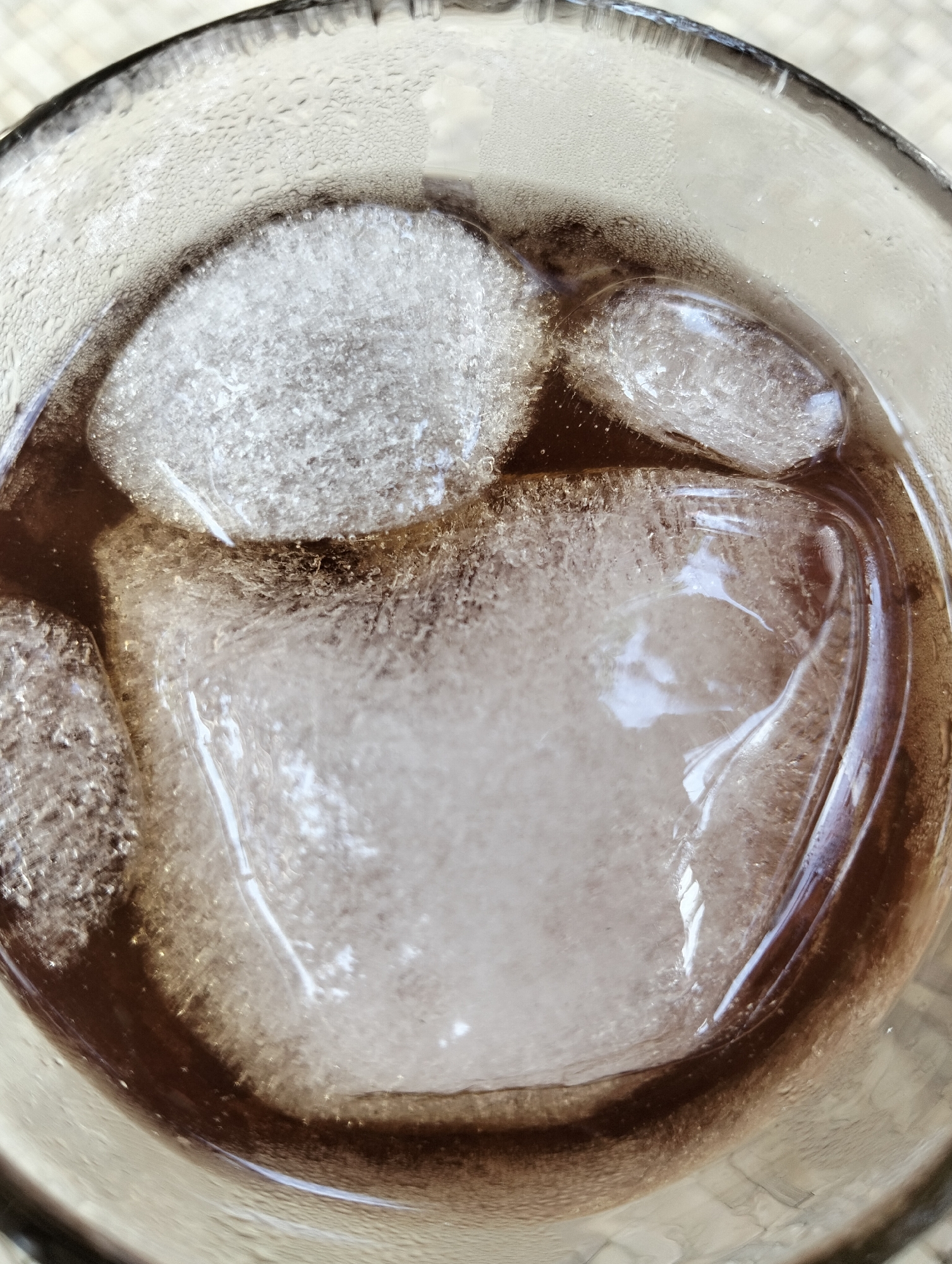 This is a close-up, top-down shot of a glass filled with a dark liquid and ice cubes. The focus is on the ice and the condensation on the outside of the glass, suggesting a cold drink. The ice cubes vary in size and shape, with one large cube dominating the center of the frame. The liquid appears to be a dark brown, possibly cola or iced coffee. The glass has condensation droplets on its exterior, indicating it's cold and likely recently filled or sitting in a warm environment. The background is blurred but appears to be a textured surface, possibly a table. There is no visible text, people, or specific activity, just the static image of a refreshing drink. The setting could be indoors or outdoors, but the presence of condensation implies a need for a cold beverage, possibly on a warm day in Cottrueng, Indonesia.