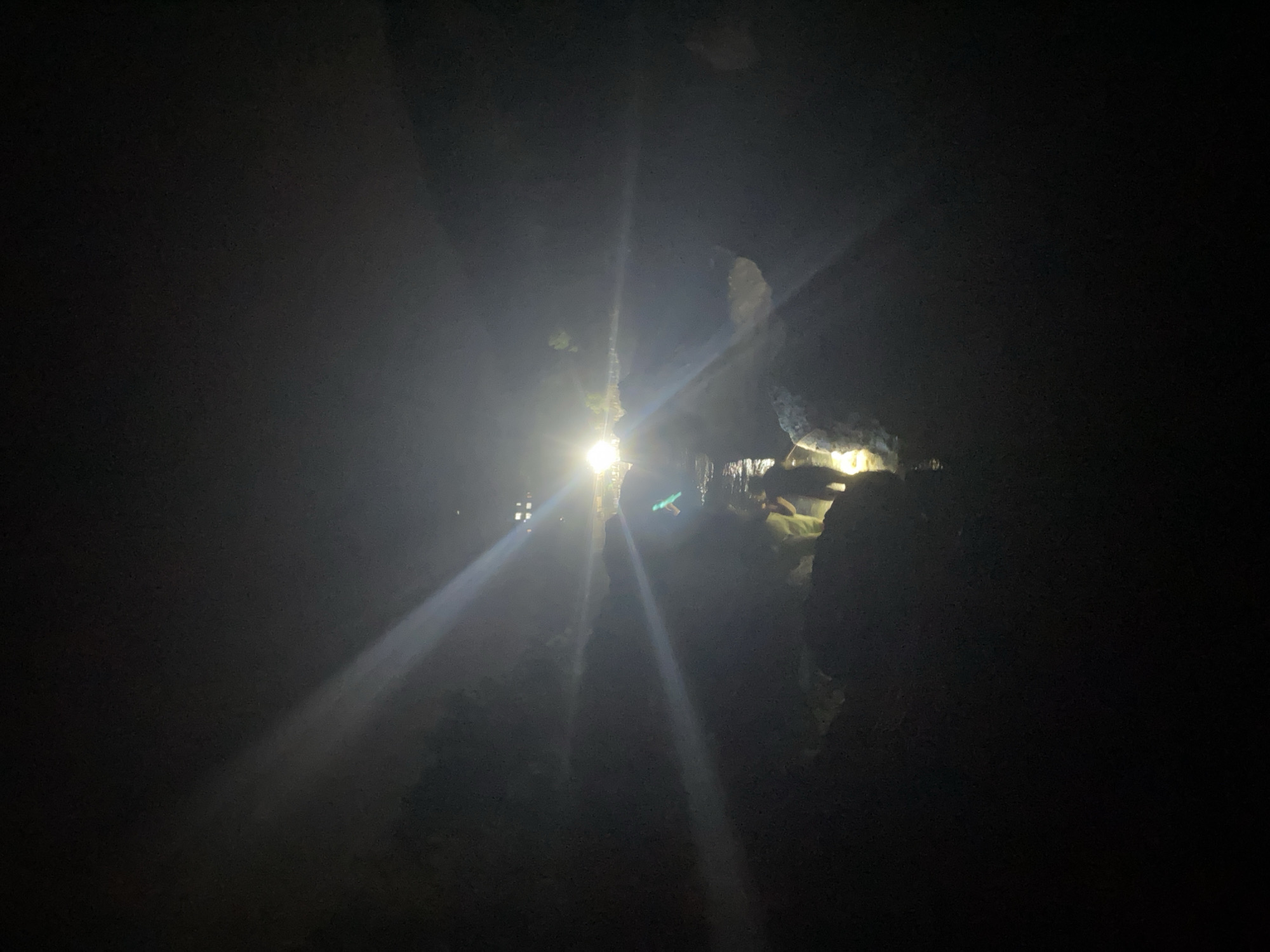 The image depicts a nighttime scene in Sutomore, Montenegro, focusing on a group of individuals navigating a rocky, shallow body of water. Bright, intense lights, possibly from floodlights or a vehicle, illuminate the area, creating strong beams that cut through the darkness and cause lens flares. Several people are visible, some wading in the water, while others stand on the rocky banks. One individual in the foreground appears to be holding a light source, casting a greenish glow. The environment is characterized by dark, rough terrain and what appears to be a riverbed or coastal area. The overall impression is of an organized activity, possibly a rescue operation or an exploration, taking place under challenging, low-light conditions.