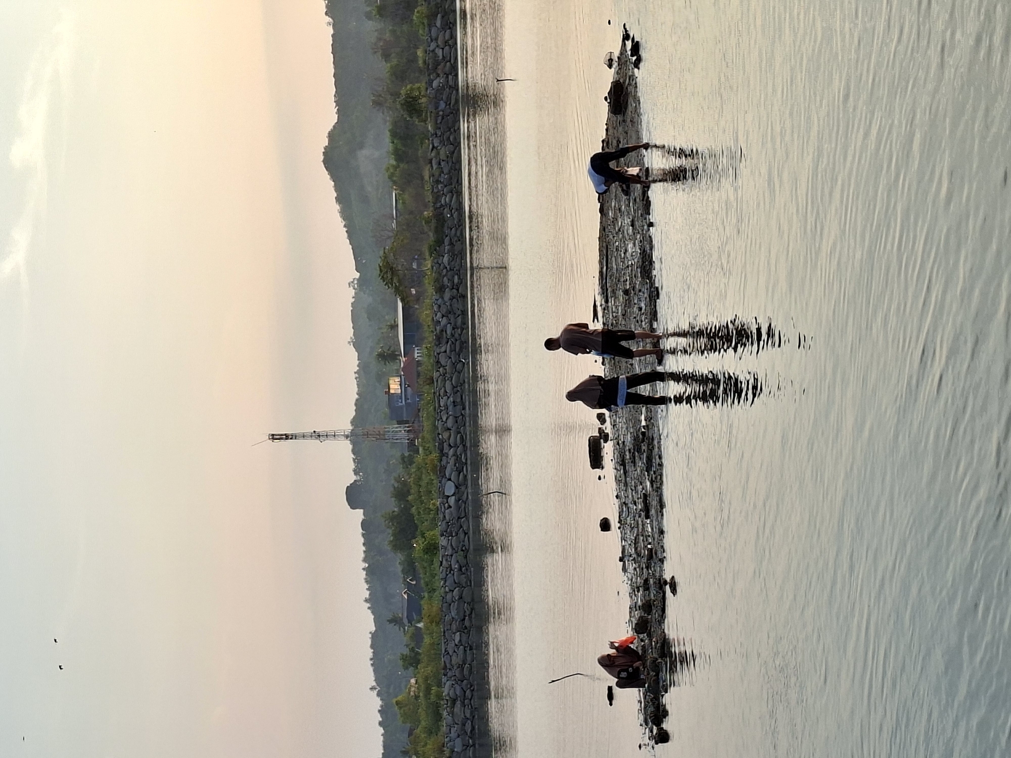 The image shows a group of people standing on a narrow strip of land extending into the water. The scene is likely in the city of Lhokseumawe, Indonesia, given the request. A line of trees and a tower can be seen along the water's edge. The sky is bright, suggesting sunrise or sunset. The water is calm, with slight ripples. The people seem to be engaged in activities such as fishing or collecting items from the shore.
