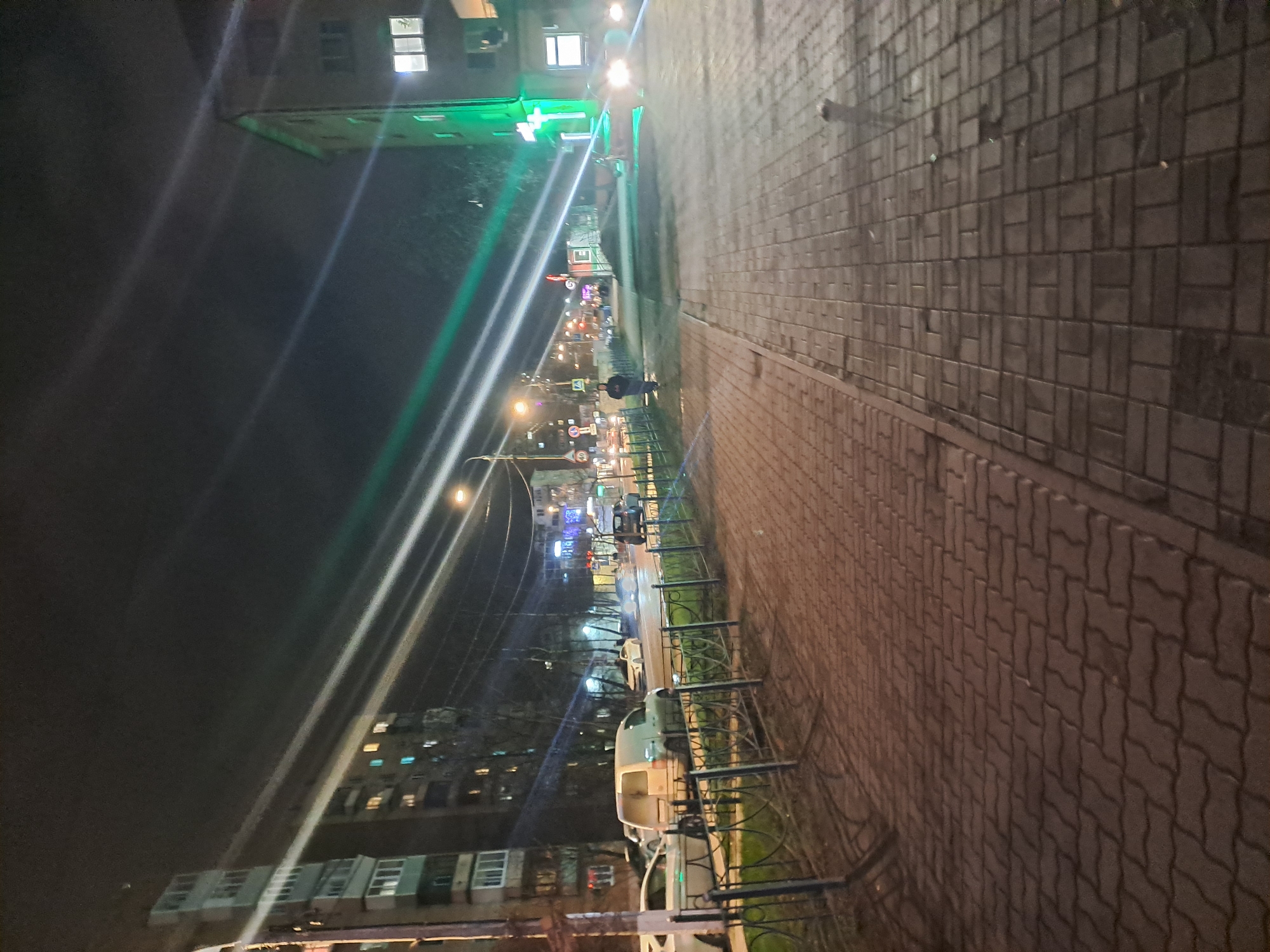 The image depicts a street scene at night in Elektrostal, Russia. The perspective is oriented such that the buildings on the left side appear vertically, while the walkway on the right is horizontal. Streetlights illuminate the scene, causing bright glare in some areas. On the left, multi-story residential buildings with lit windows are visible. Some buildings feature green lighting. A street stretches into the distance, with various traffic lights, signs, and vehicles. A sidewalk runs along the right side of the street. A low metal fence separates the sidewalk from the road. The sidewalk is paved with interlocking pavers. A person is standing near the edge of the sidewalk.