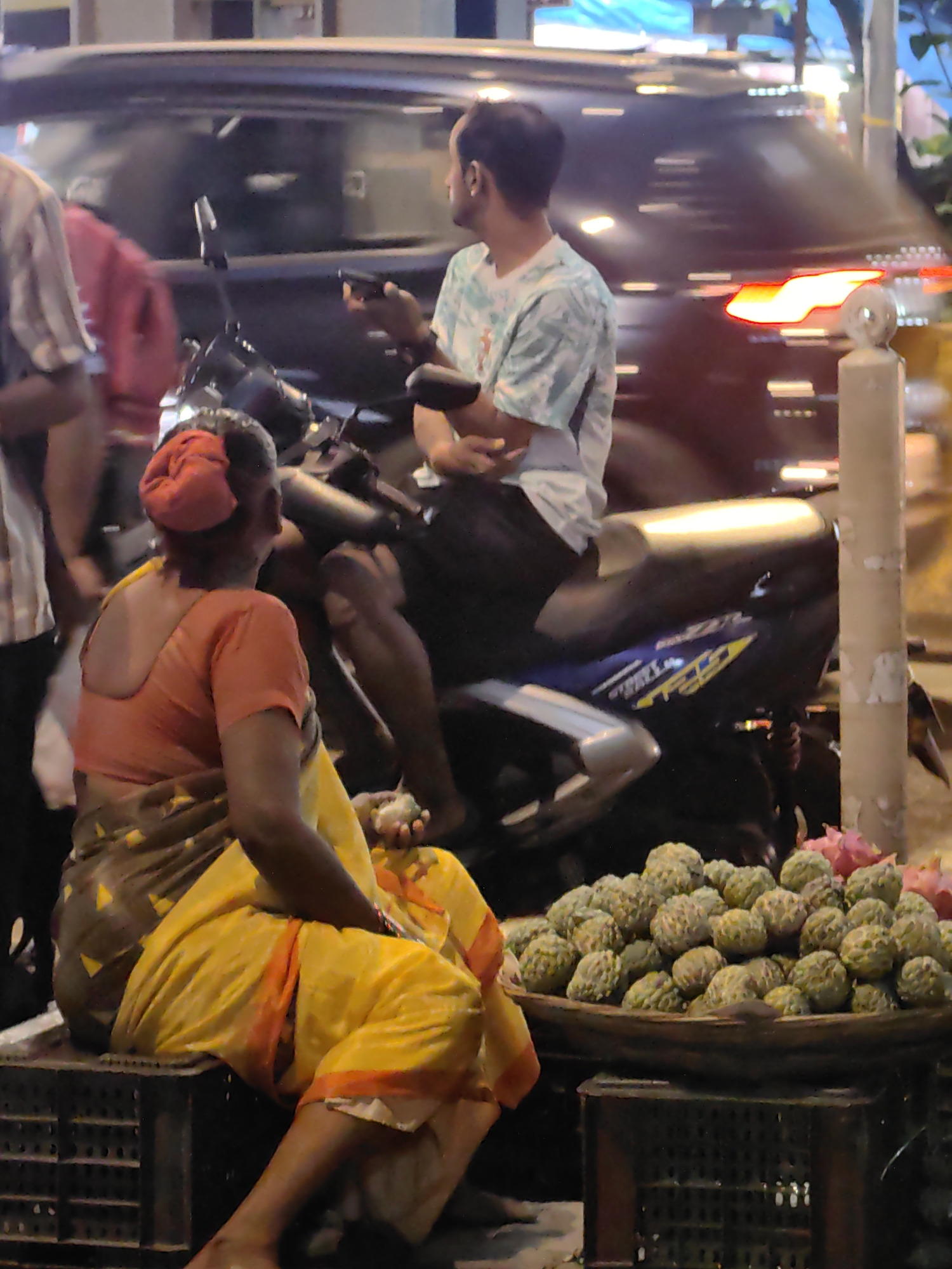 In Mira Bhayandar, India, a woman in a traditional sari sits on a crate, selling fruit from a basket. She wears an orange blouse and a yellow sari, with her hair tied up in a red cloth. In front of her is a large basket overflowing with custard apples, and a few pink dragon fruits are visible at the back of the basket. To her right, a man on a scooter looks at his phone. In the background, a dark-colored car is visible, with its taillights illuminated. The scene appears to be a street at dusk or night, with artificial lighting creating reflections on the car and illuminating the fruits. The overall impression is of a busy street scene in an Indian city.