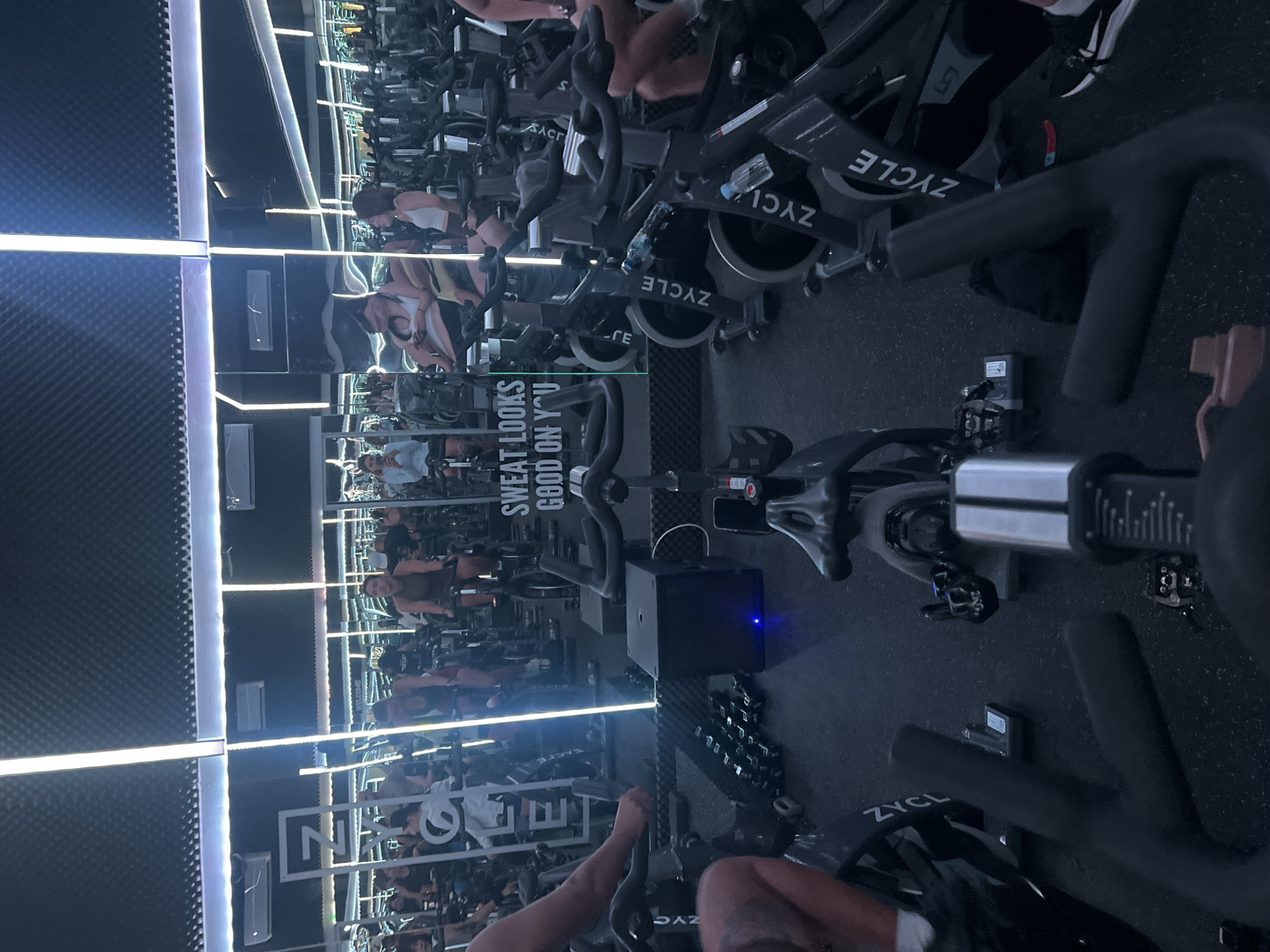 This image captures a fitness studio with rows of stationary bicycles, likely a spin class. The environment is dimly lit, with bright, vertical LED lights illuminating the space and reflecting off mirrored walls, creating a dynamic and energetic atmosphere. In the foreground, several exercise bikes are visible, some with water bottles attached. The word "ZYCLE" is prominently displayed on the side of several bicycles. In the background, multiple individuals are seated on bicycles, engaged in exercise. A sign on the mirrored wall reads "SWEAT LOOKS GOOD ON YOU." The overall impression is that of a modern, high-energy fitness facility.