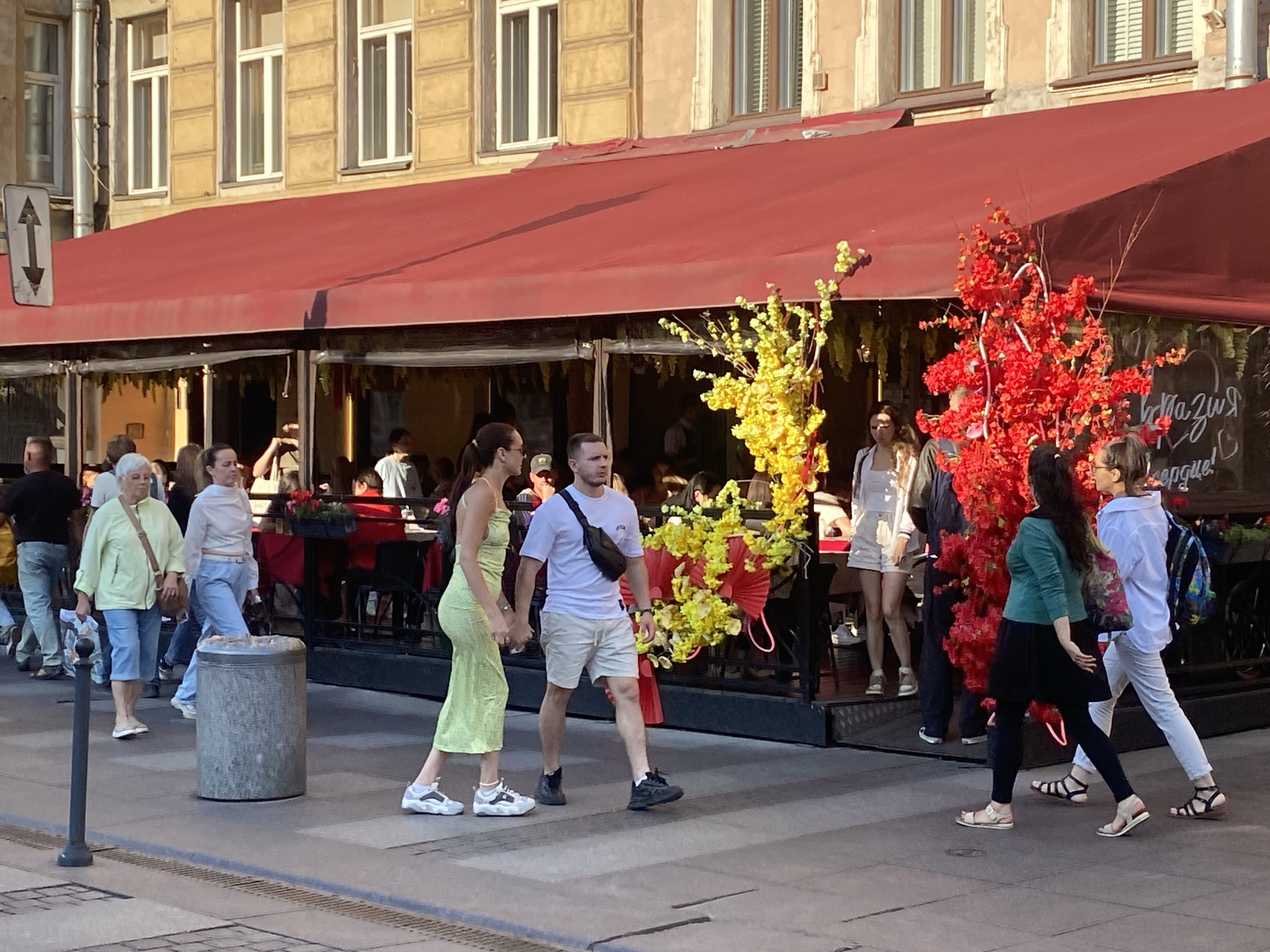 The image depicts a street scene in Saint Petersburg, Russia, with a building facade and a red awning of a cafe or restaurant dominating the background. Several people are walking on the sidewalk in front of the establishment. On the left, a woman in a light green dress and a man in a white t-shirt and beige shorts are walking hand-in-hand. To their right, a woman in a light blue shirt and jeans walks past a cylindrical trash bin. Further to the right, a woman in a green top and black leggings walks with a backpack, followed by a woman in a white shirt and white pants. Decorative artificial trees with red and yellow blossoms are placed in front of the cafe, adding a touch of color to the scene. The sidewalk appears to be made of paving stones.