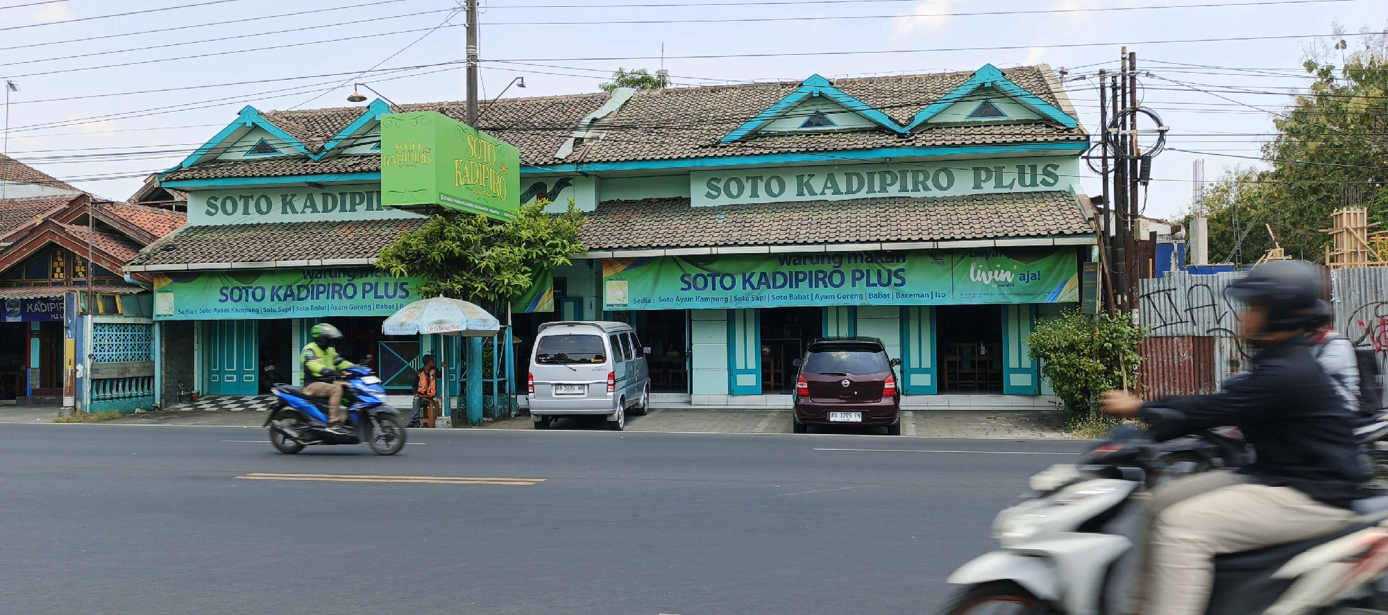 The image captures a street view of "Soto Kadipiro Plus," a restaurant located in Kasihan, Indonesia. The building, painted in light turquoise and white, features a tiled, mansard-style roof with decorative triangular gables. Large green banners with the restaurant's name and menu items are prominently displayed on the facade. In front of the restaurant, a silver van is parked, and a motorcycle with a rider in a bright green jacket is passing by. Further down the street, another motorcycle rider, with their face blurred due to motion, is visible. The environment includes a utility pole with a complex network of wires, a construction site with scaffolding, and trees. The sky is overcast.