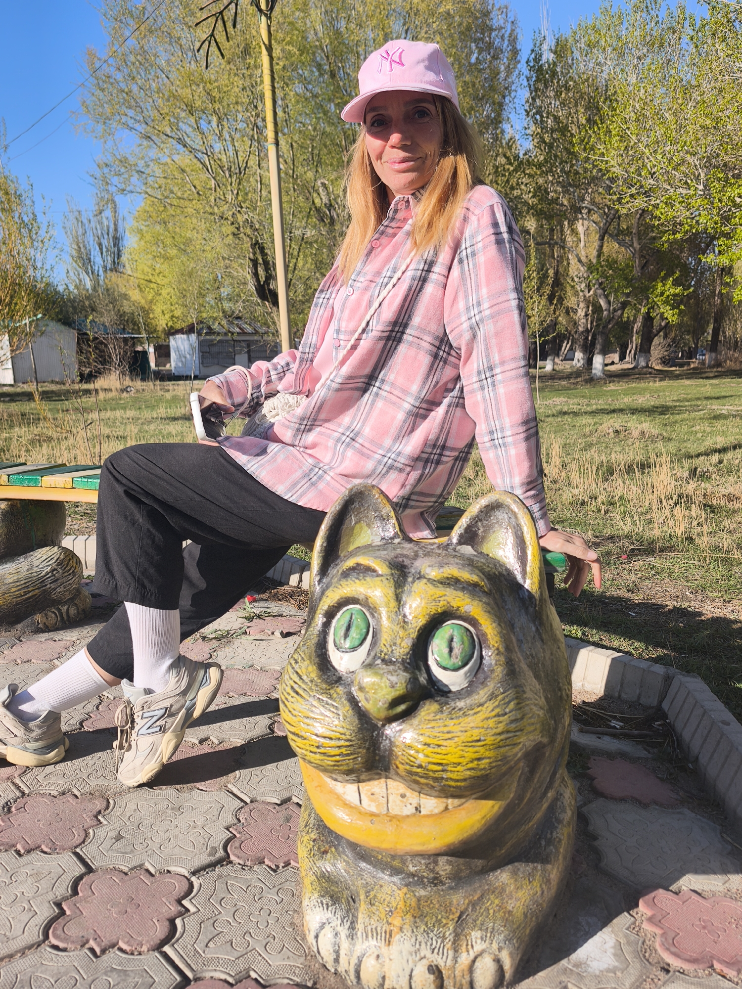 A woman sits on a green park bench next to a painted concrete statue of a smiling cat. The woman wears a pink baseball cap with a white logo, a pink and gray plaid flannel shirt over a lighter pink shirt, black pants cuffed above the ankles, white socks, and beige New Balance sneakers. She holds a white smartphone in her left hand and rests her right hand on the bench. The cat statue is painted yellow, gray, and black with large, green eyes and a wide, toothy grin. The ground is covered in red and gray paving stones. Trees are visible in the background. The location is Balykchy, Kyrgyzstan.