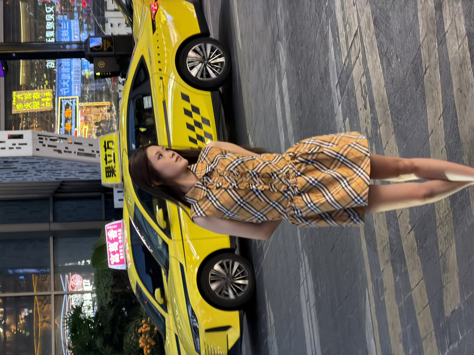 In Yuzhong District, China, a young woman in a plaid sleeveless dress stands on a pedestrian crossing at night. She is facing to her left, looking away from the camera, with her hands clasped behind her back. Behind her, two yellow taxis are parked side-by-side. The taxi on the left has a pink illuminated sign on its roof with Chinese characters. The taxi on the right has a yellow illuminated sign with Chinese characters that read "果立方". The background features brightly lit buildings with neon signs. The street is paved with dark asphalt and marked with yellow and grey pedestrian crossing stripes. The lighting suggests it is evening or night.
