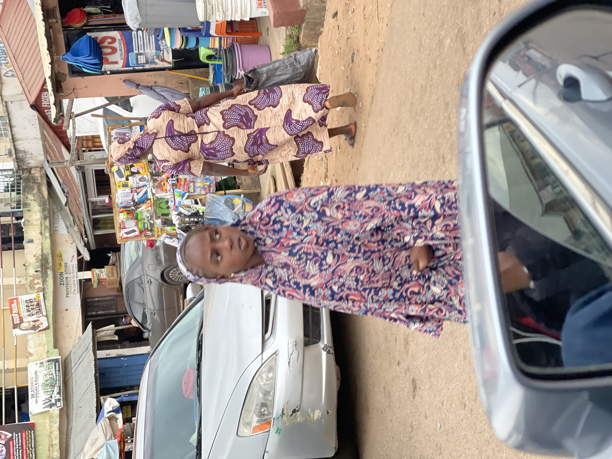 The image captures a street scene, likely in the city of Jos, Nigeria. Two individuals, both women, are featured. One woman, wearing a patterned, beige and purple dress, stands in the background, while the second woman, wearing a patterned, blue and red dress, is positioned closer to the camera, next to a silver car with visible damage to the front bumper.
The scene unfolds on a paved road in a commercial area. In the background, a shop front displays a variety of goods. A signboard reads "ZION PROVISION STORE" and other adjacent signs advertise "SK STORES," "WINE, BEER AND SOFT DRINKS" and "FABILOUS DEZ FROZEN POS AVAILABLE HERE." Another silver car is parked behind the damaged one. The side mirror of the car closest to the viewer occupies a portion of the right side of the frame. The lighting suggests it's daytime.