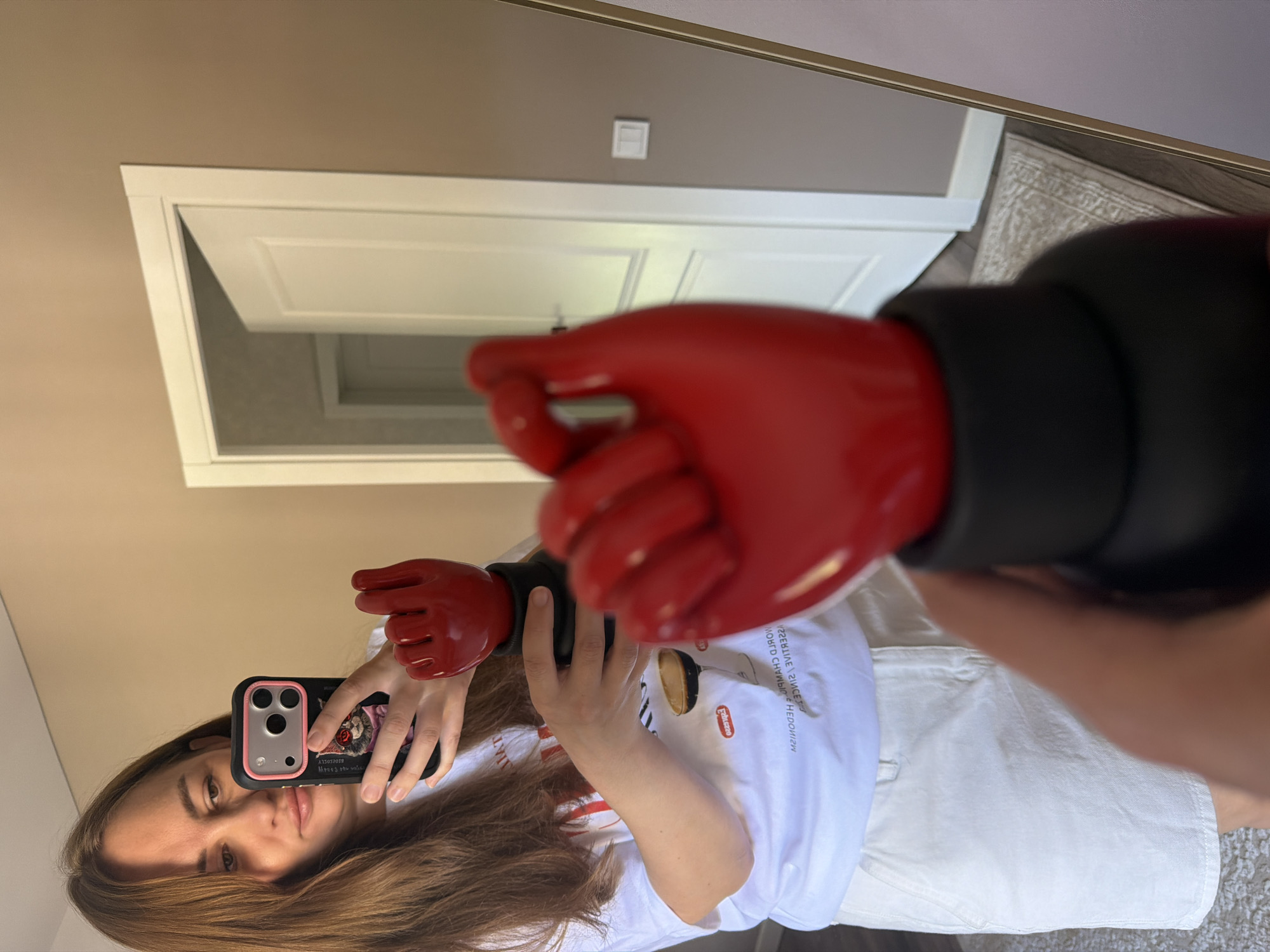 A woman is taking a mirror selfie in what appears to be a home interior. She is holding a smartphone in her right hand and a red fist-shaped object in her left. She is wearing a white t-shirt and a white skirt. In the background, a white door is ajar. The environment is neutral, with beige walls and a wooden floor visible in the reflection. The overall impression is casual and domestic. The image does not provide any information about the city of Malinovka, Russia.