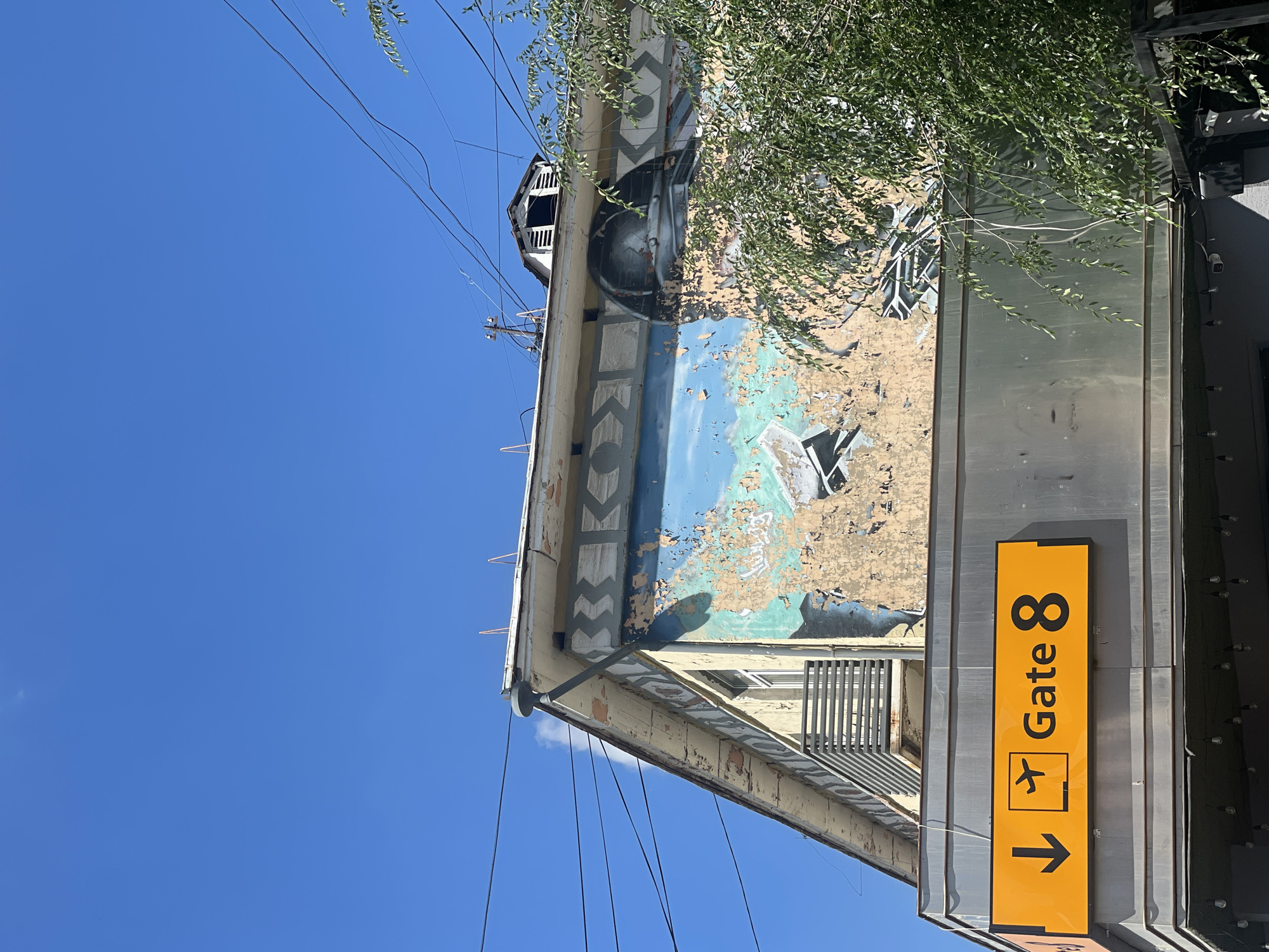 This image captures a section of a building's exterior under a clear blue sky. Prominently displayed is a bright orange sign with black lettering and symbols that reads "Gate 8," accompanied by an airplane icon and a downward-pointing arrow. Above the sign, the building facade is weathered, with peeling paint revealing layers underneath. A mural, partially obscured by the peeling paint and a leafy green tree, is visible on the upper part of the building. The mural appears to depict a large, metallic-domed object, possibly a helmet or a spacecraft, against a blue background. Geometric patterns are also visible along the upper edge of the building. Several overhead wires crisscross the sky. The overall scene suggests an urban setting, possibly near an airport or a transportation hub, but the specific location in Ulaanbaatar, Mongolia, cannot be determined from the visual information alone. There are no people visible in the image.