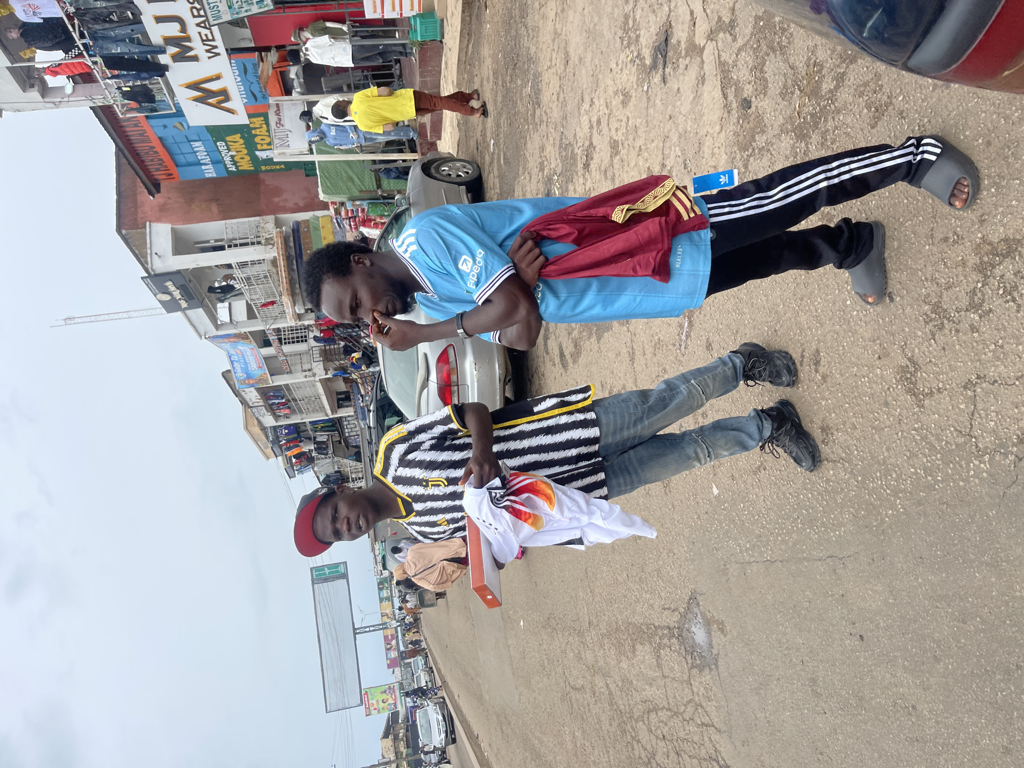 Two young Black men are standing on a paved road in Jos, Nigeria. The man on the left wears a black and white striped jersey with yellow accents, a red baseball cap, blue jeans, and black sneakers. He holds a shoebox in his left hand and a white shirt with red and yellow accents in his right. The man on the right wears a light blue short-sleeved t-shirt with white and black trim, black Adidas track pants with white stripes, and gray slides. He holds a dark red garment with gold trim in his right hand and is touching his nose with his left hand.
Behind them, storefronts line the street, with clothes and other merchandise displayed on racks and mannequins. A silver sedan is parked on the right side of the road, and other vehicles are visible in the background. The sky is overcast, and the ground is a mixture of asphalt and dirt with some puddles. The overall scene depicts a busy commercial area in a Nigerian city.